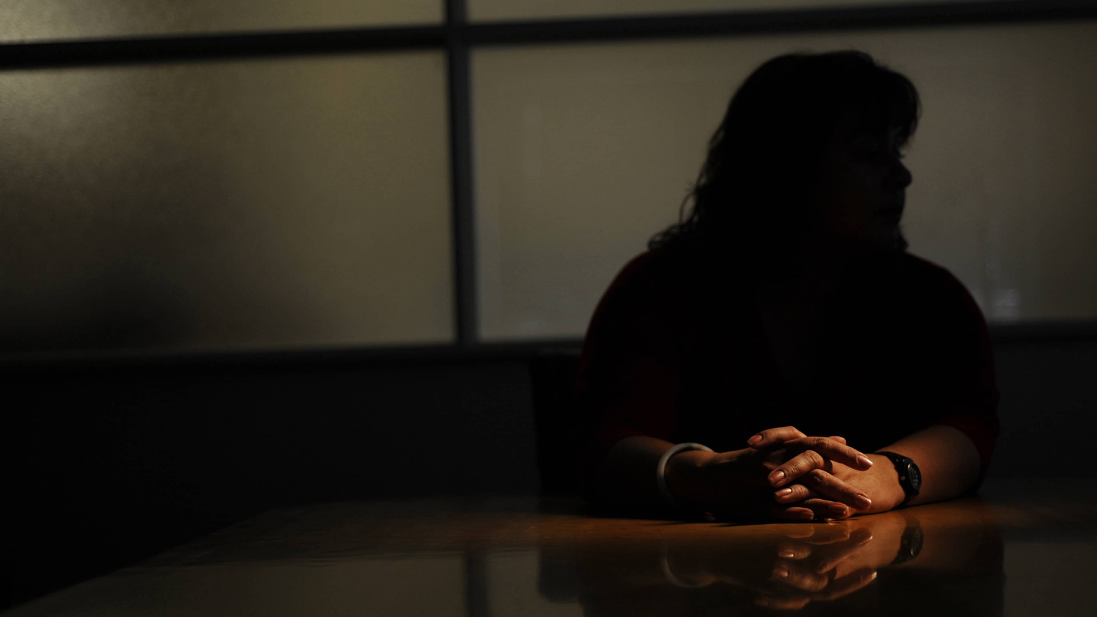 A shadowy figure stands in the background, while a person's hands are visible in the foreground, resting on a reflective surface.