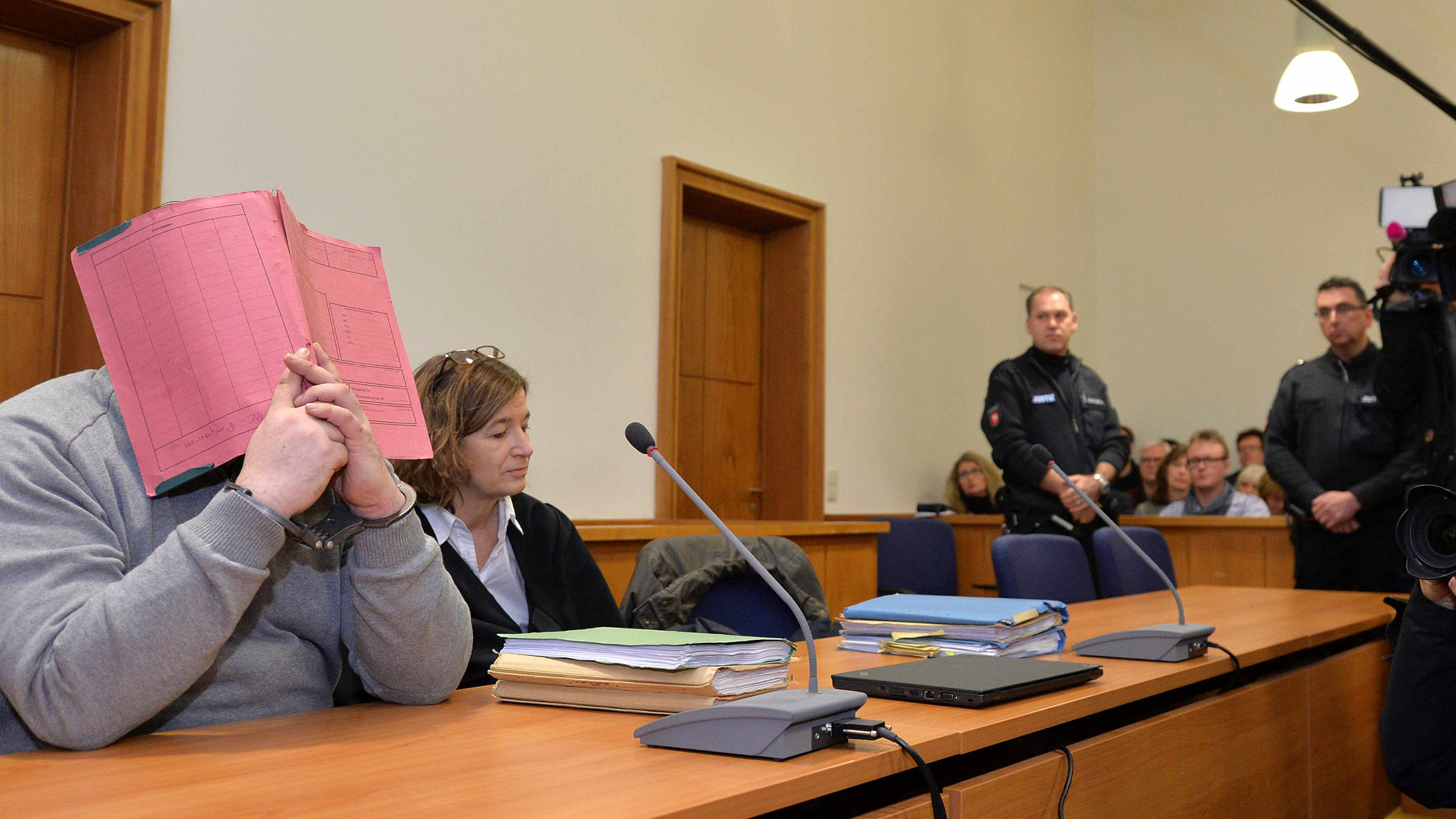 A woman sits at a table in a courtroom, holding a pink folder in front of her face, while several people, including law enforcement officers, are present in the background.