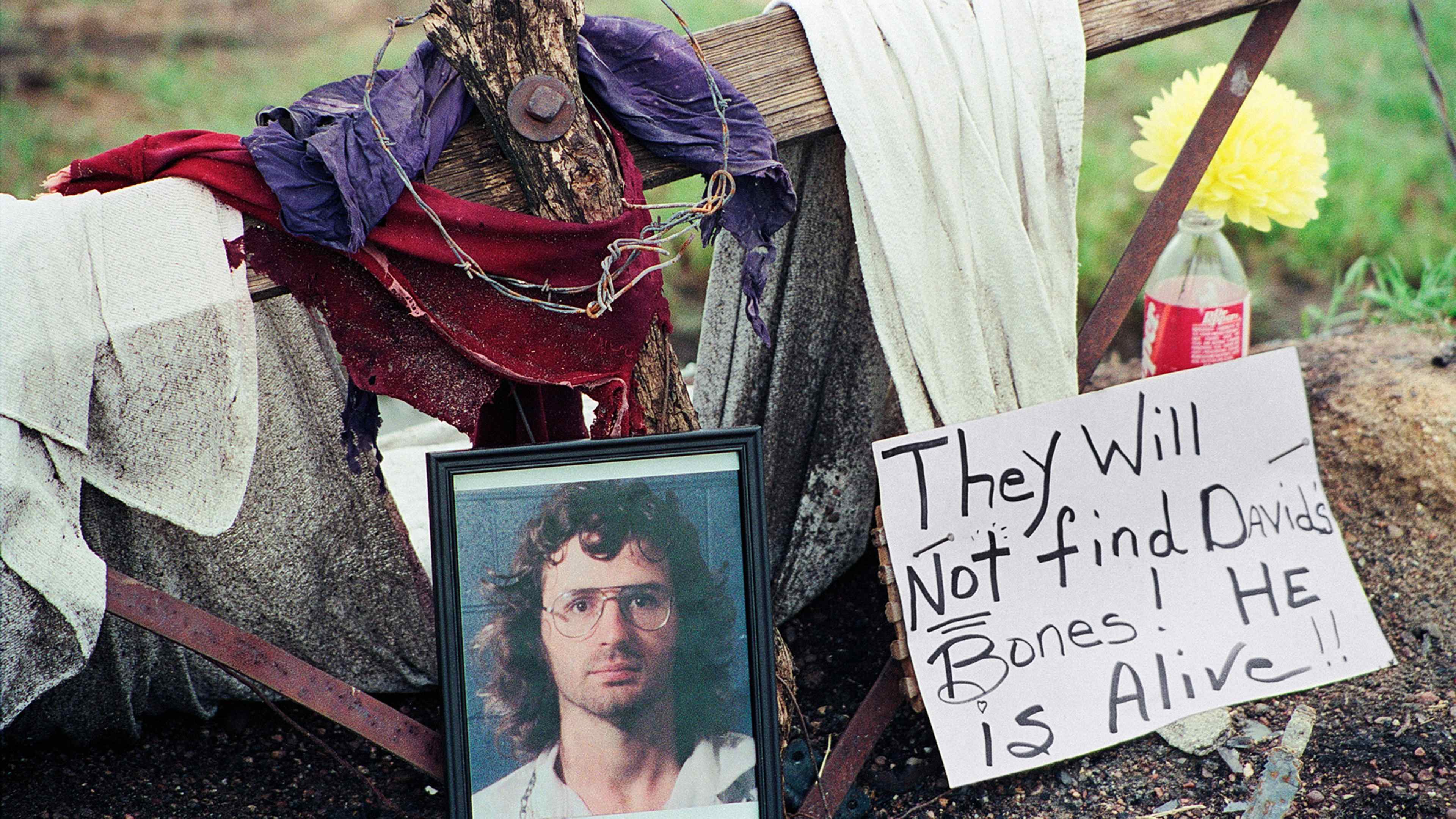 The image depicts a framed photograph of a man's face, surrounded by various items such as a red cloth, a chain, and a sign that reads "They will not find. He is alive." The background includes a wooden fence and a grassy area.