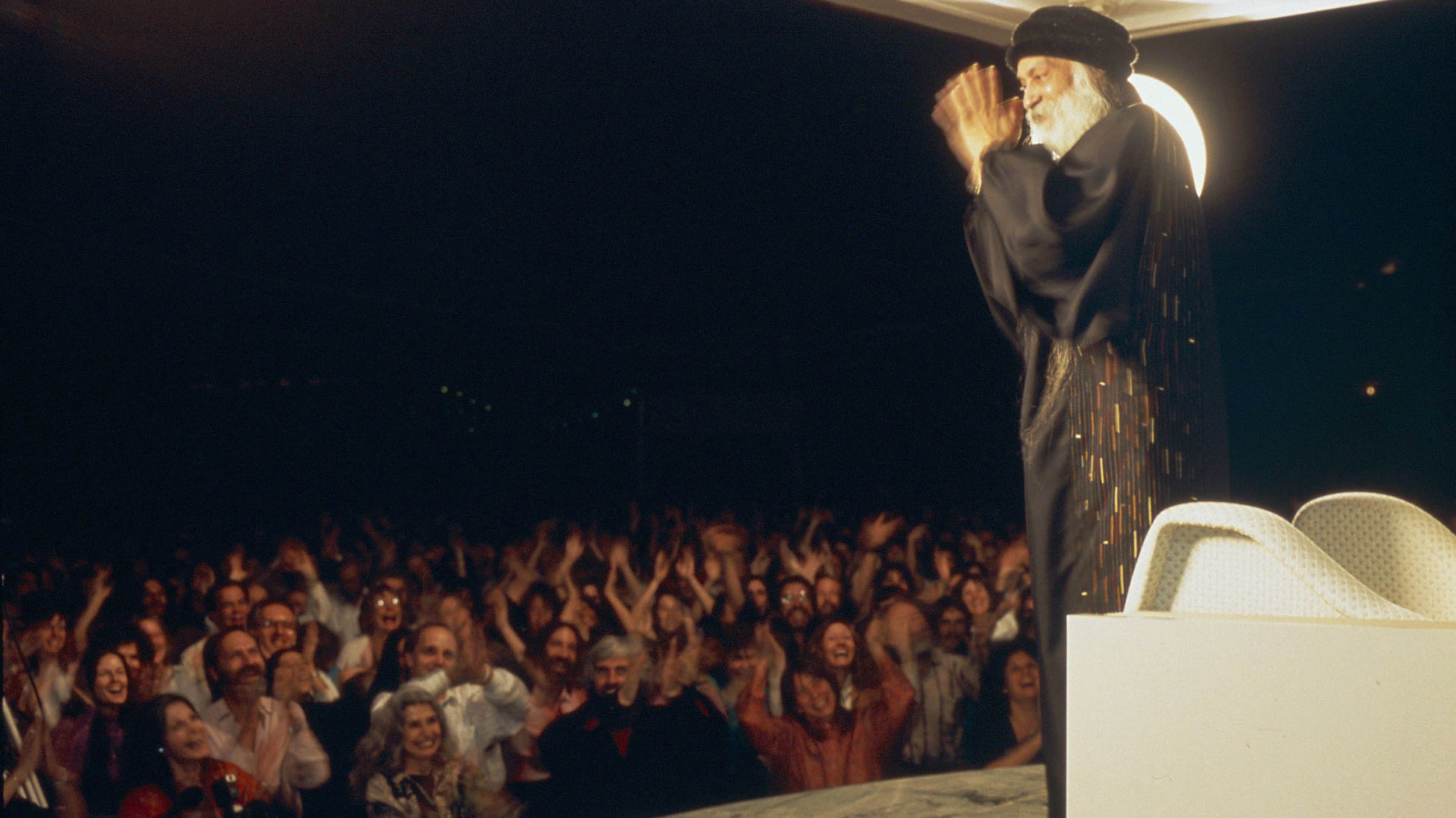 A large crowd of people with their hands raised, gathered in a dark venue, with a figure in the background appearing to be a performer or speaker on a stage.