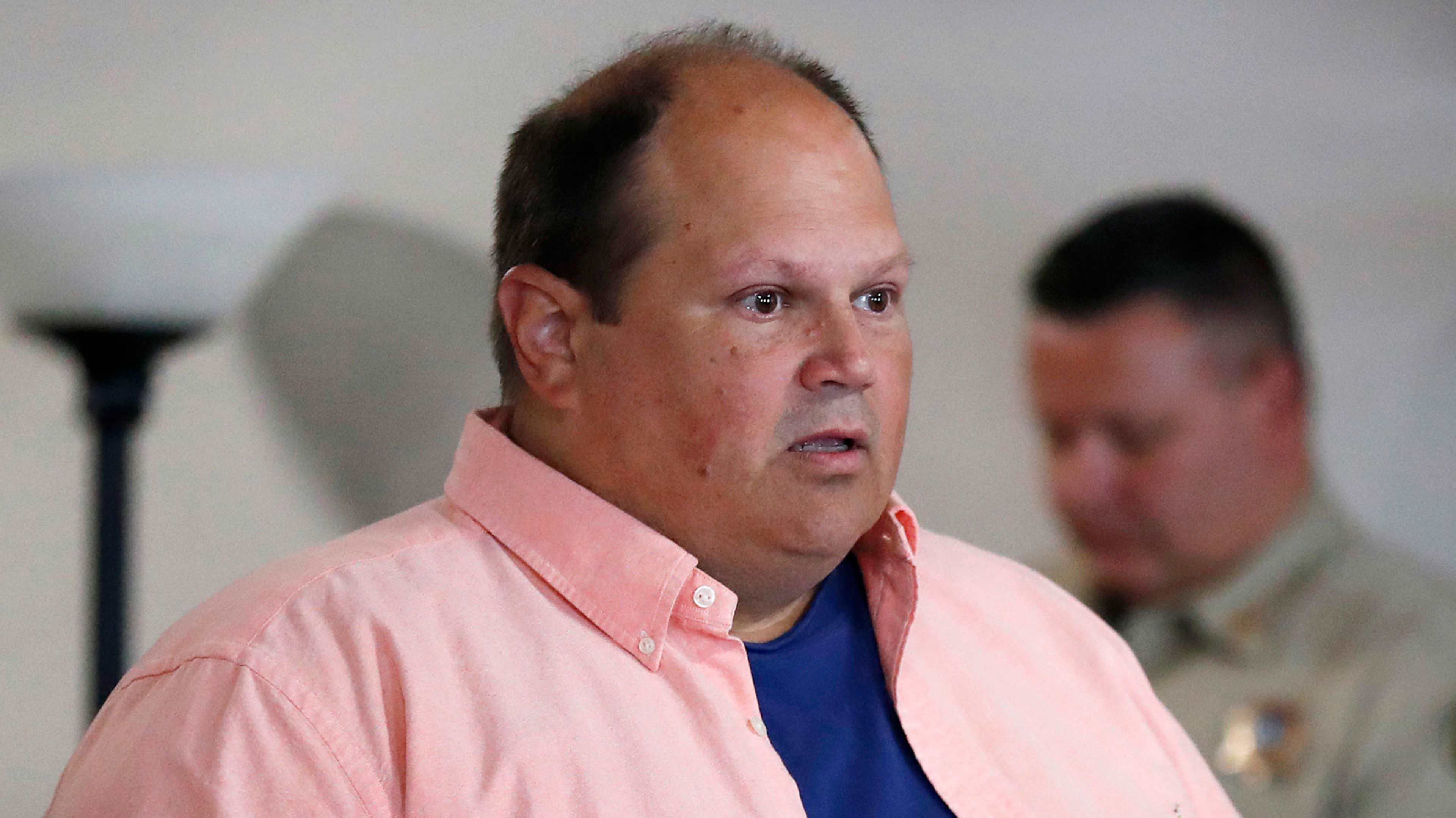 The image shows a middle-aged man wearing a pink shirt, with a serious expression on his face, standing in what appears to be an indoor setting.