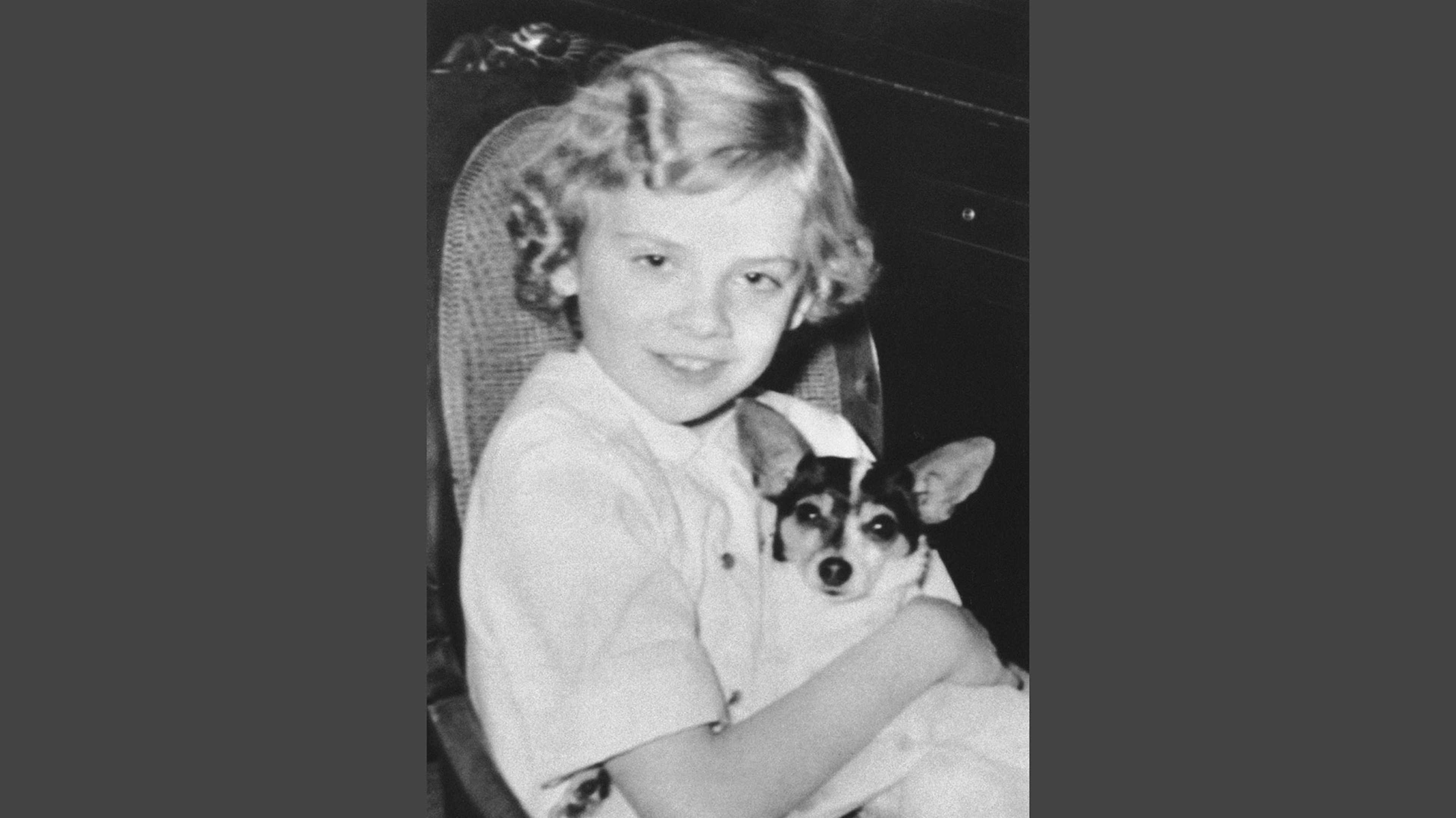 A smiling young woman with blonde curly hair is holding a small dog in her arms against a dark background.