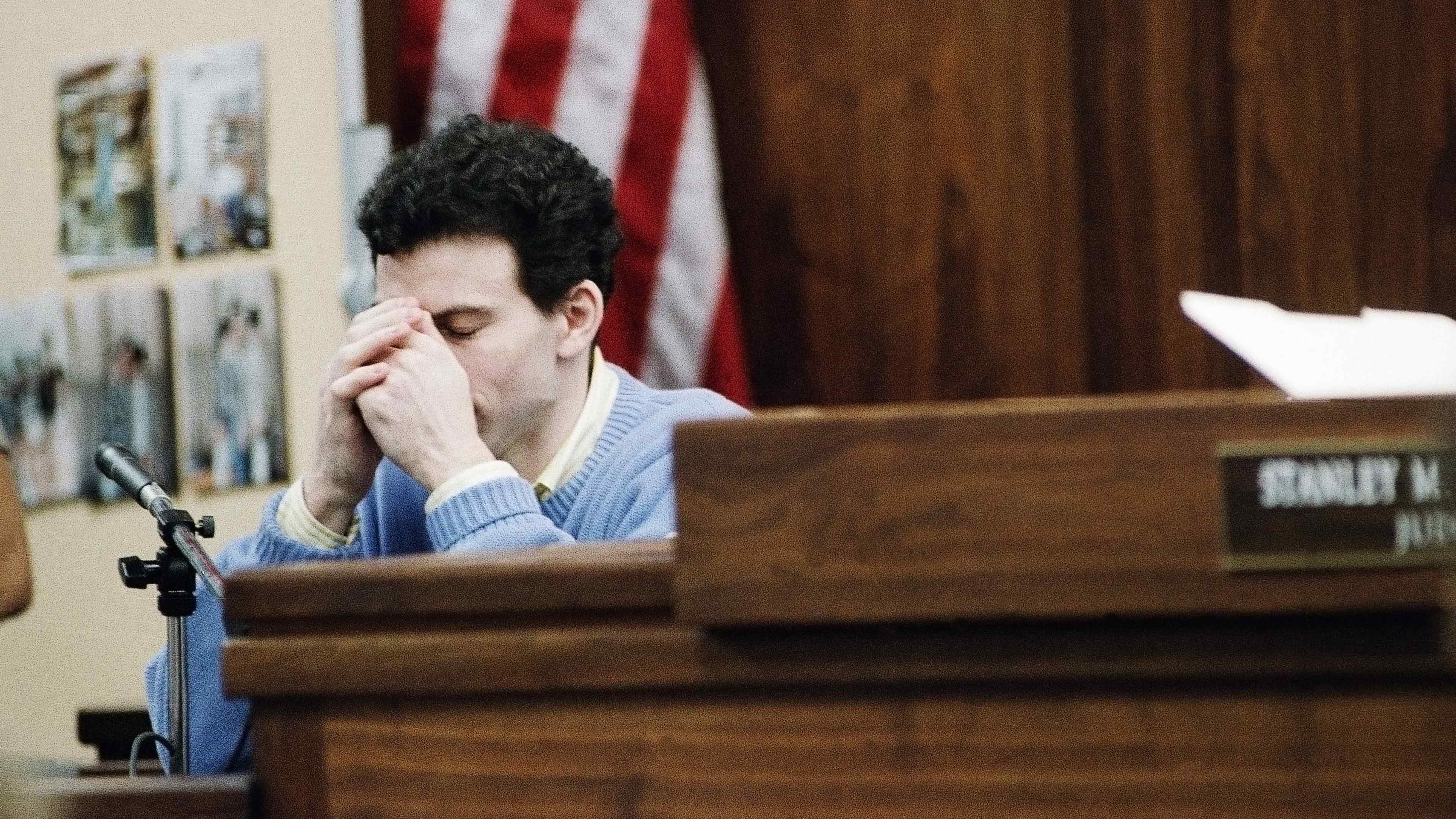 A person with dark hair is sitting at a wooden desk, with an American flag visible in the background, appearing to be in a courtroom or similar setting.