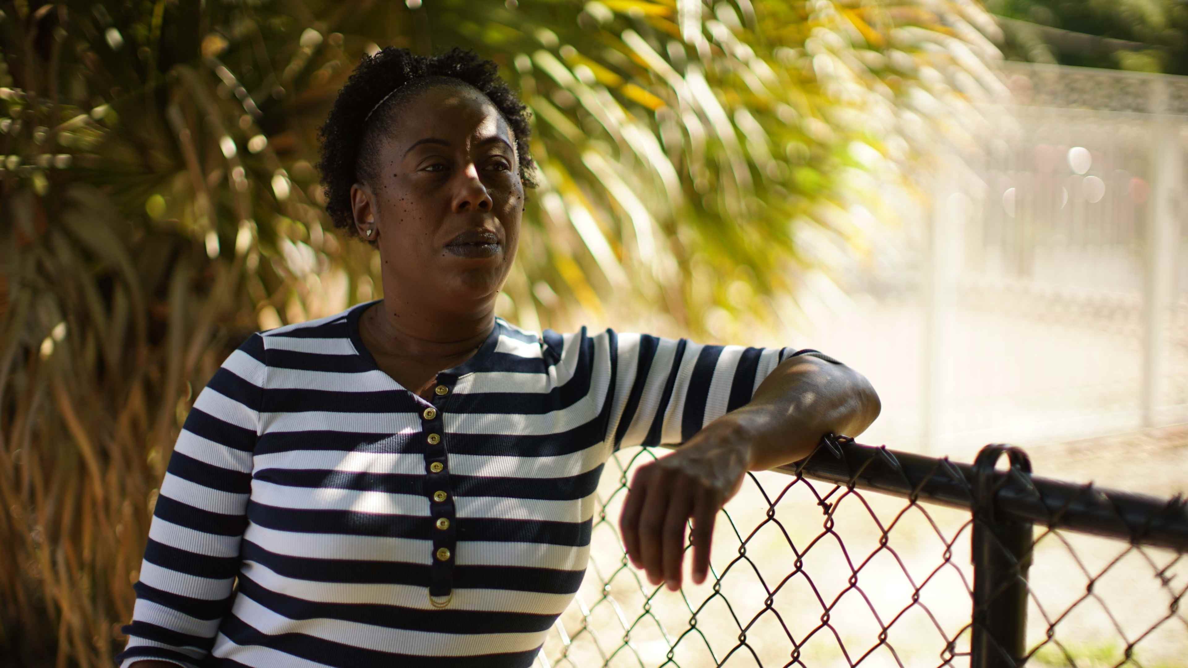 A person wearing a striped shirt stands in front of a lush, green background with palm leaves.