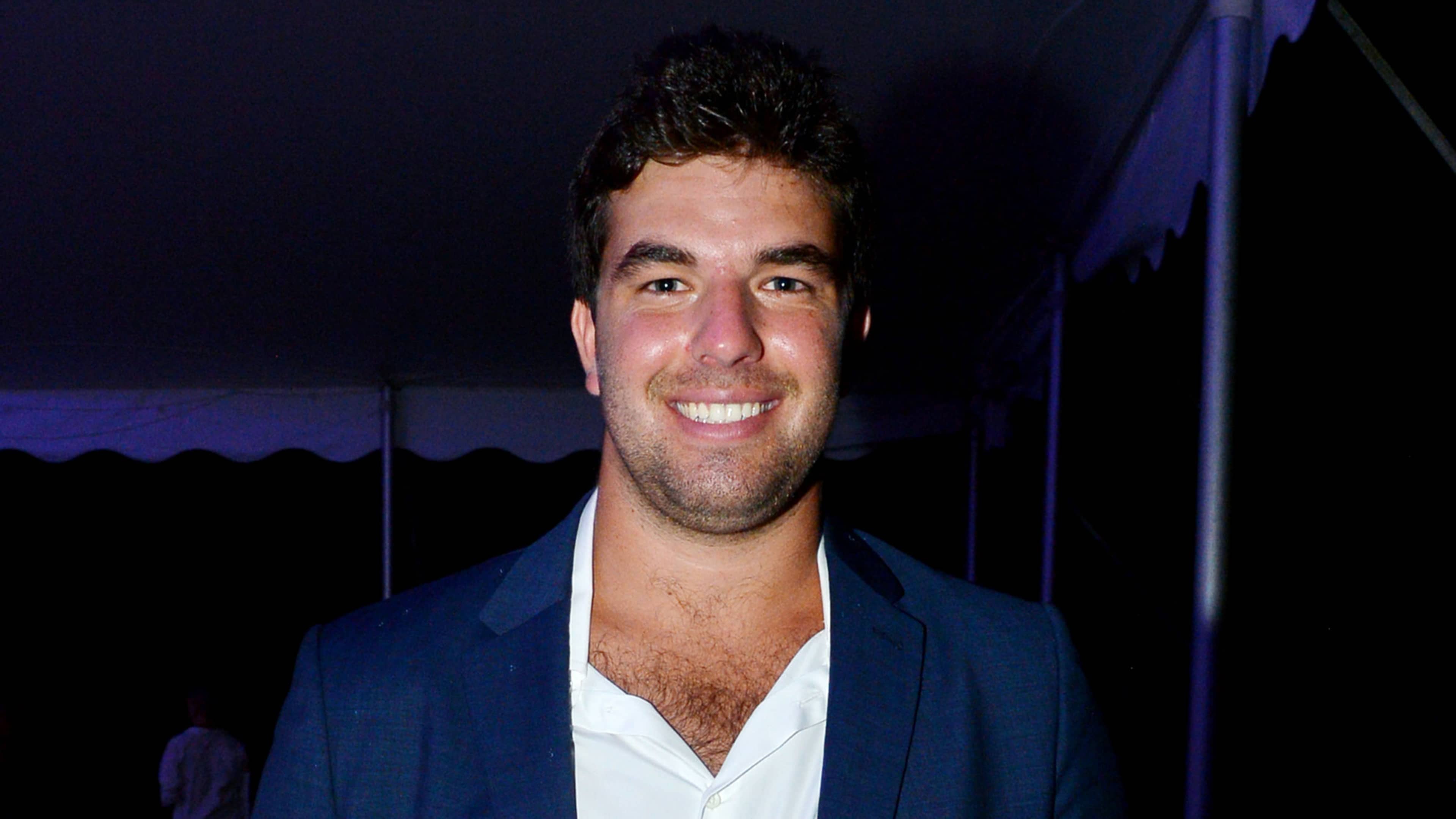 A smiling man with a beard wearing a dark blue jacket stands in front of a dark background with a purple curtain or backdrop.