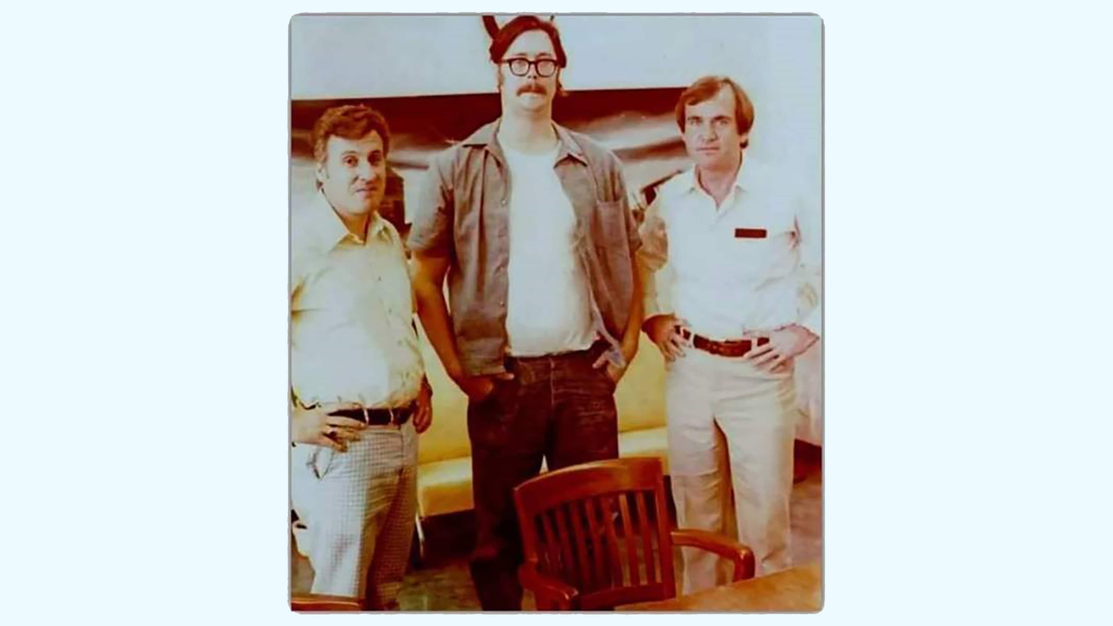 The image shows three men standing together, dressed in casual attire from what appears to be an earlier era. They have a relaxed, friendly demeanor and are positioned in front of a plain background.