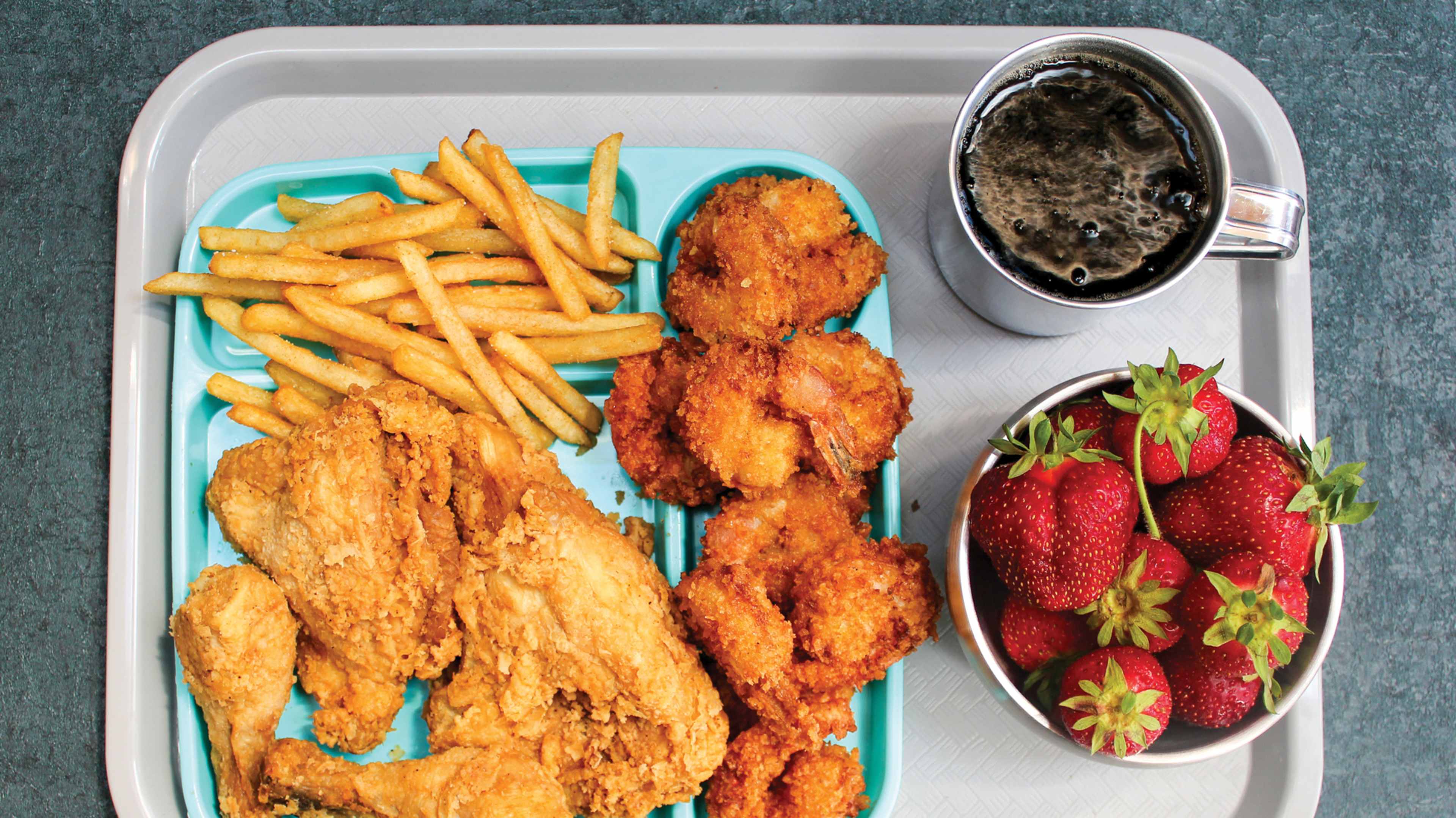 The image shows a tray with various food items, including crispy fried chicken, golden french fries, and a bowl of fresh strawberries, set against a plain background.