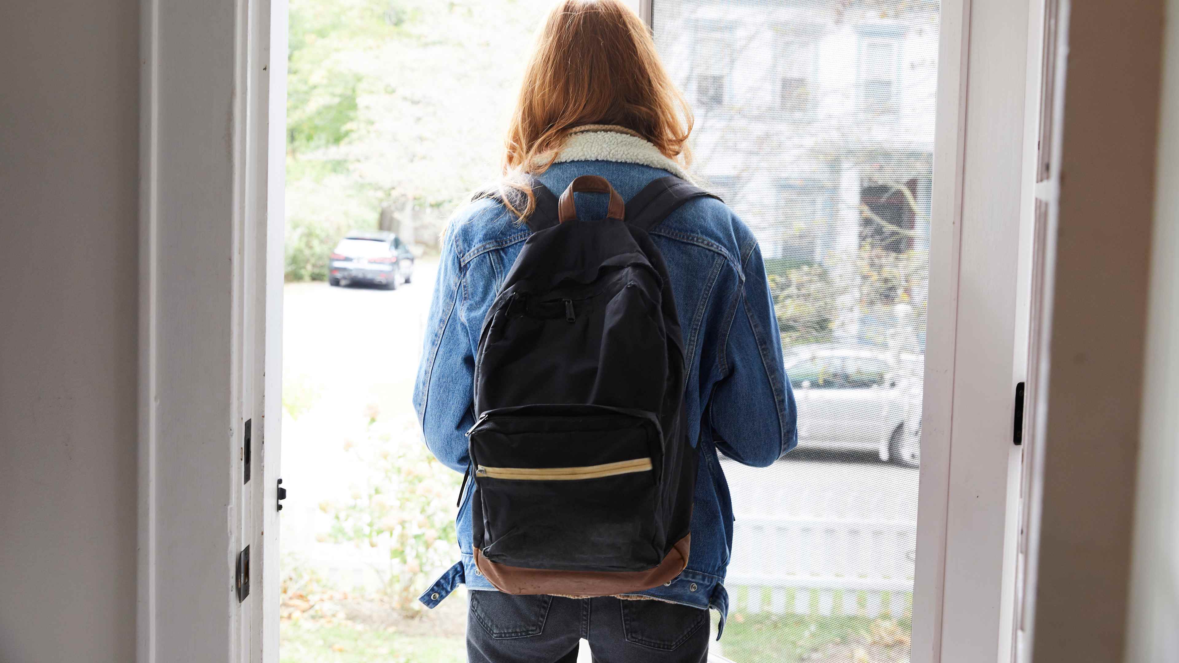 Teenage girl walking out the front door of her house. Back view of her leaving the house. She is on her way to school, wearing a back pack and holding the door open.