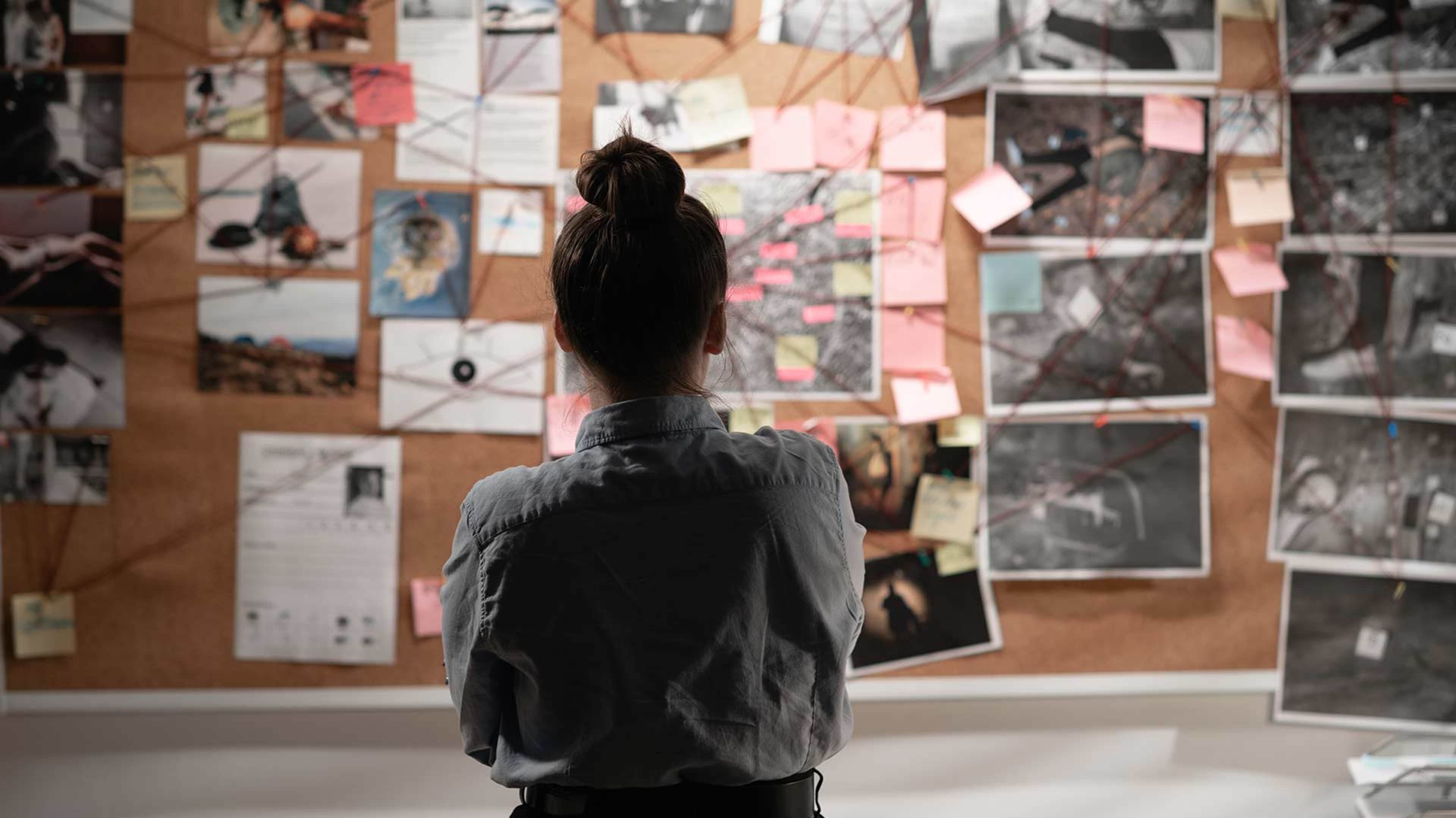 A person in a dark jacket stands in front of a wall covered with various photographs and notes.