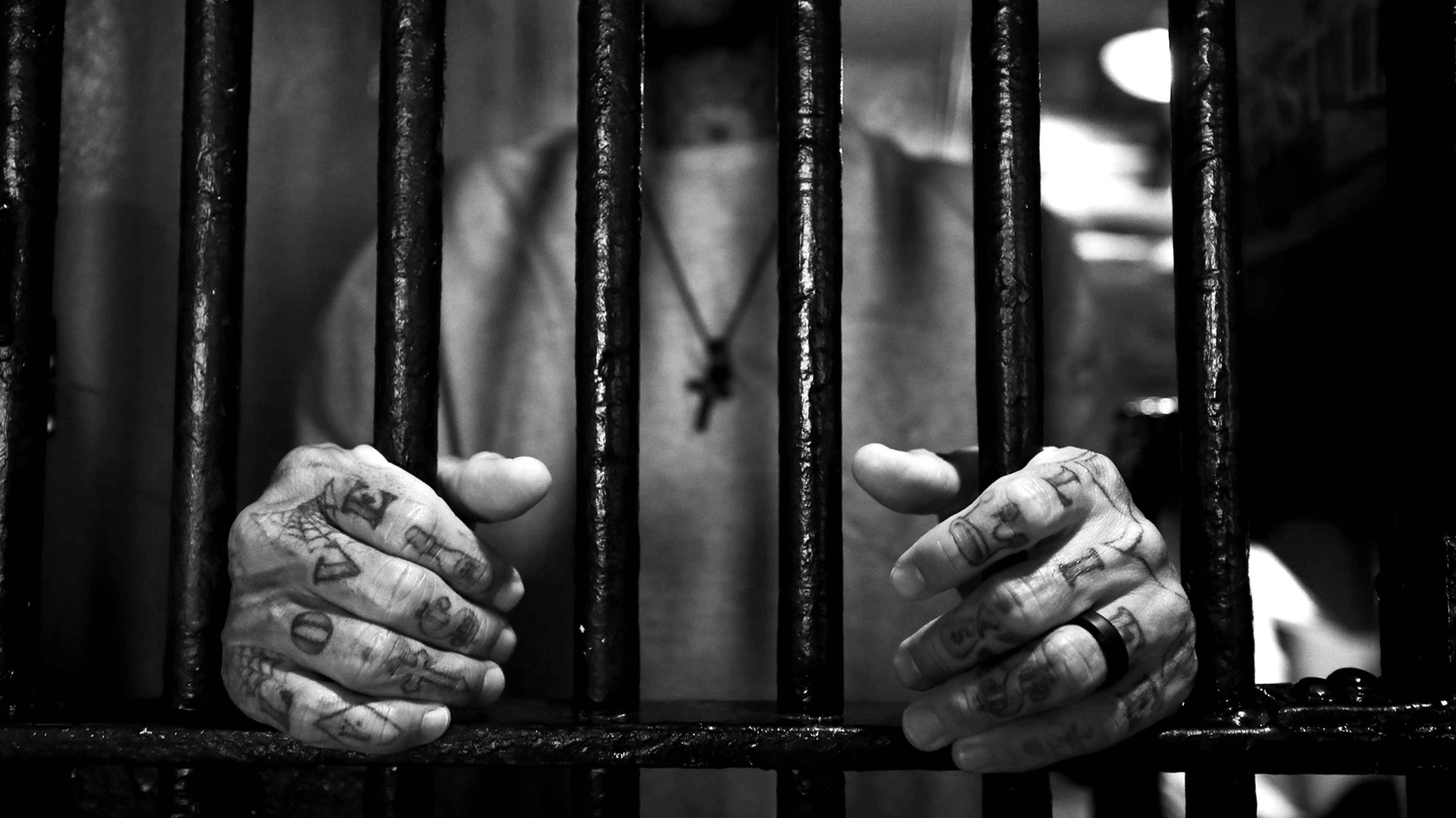 Hands grasping the bars of a prison cell, with a blurred figure visible in the background.