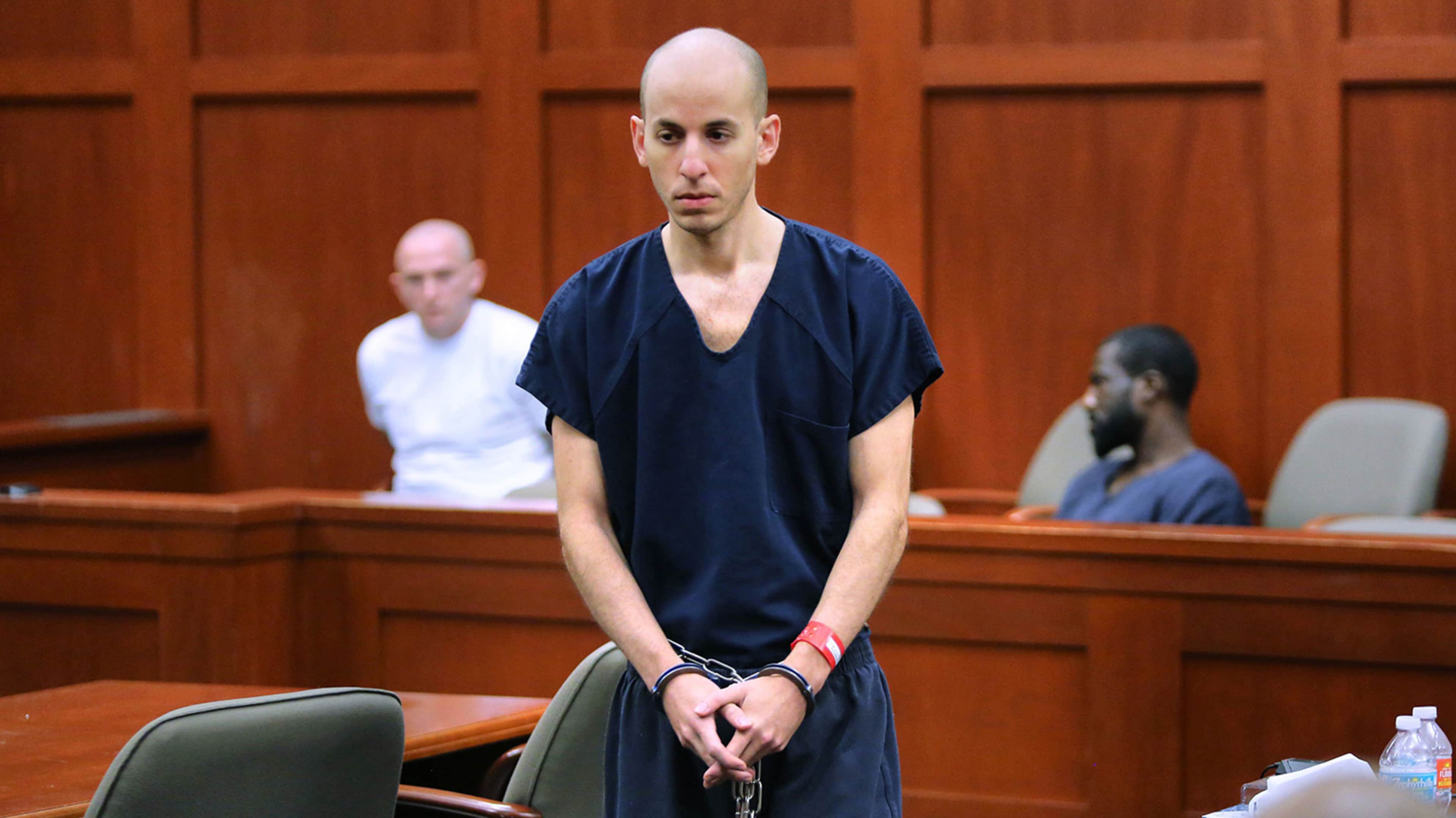 The image shows a man in a navy blue shirt standing in a courtroom, with two other individuals visible in the background.