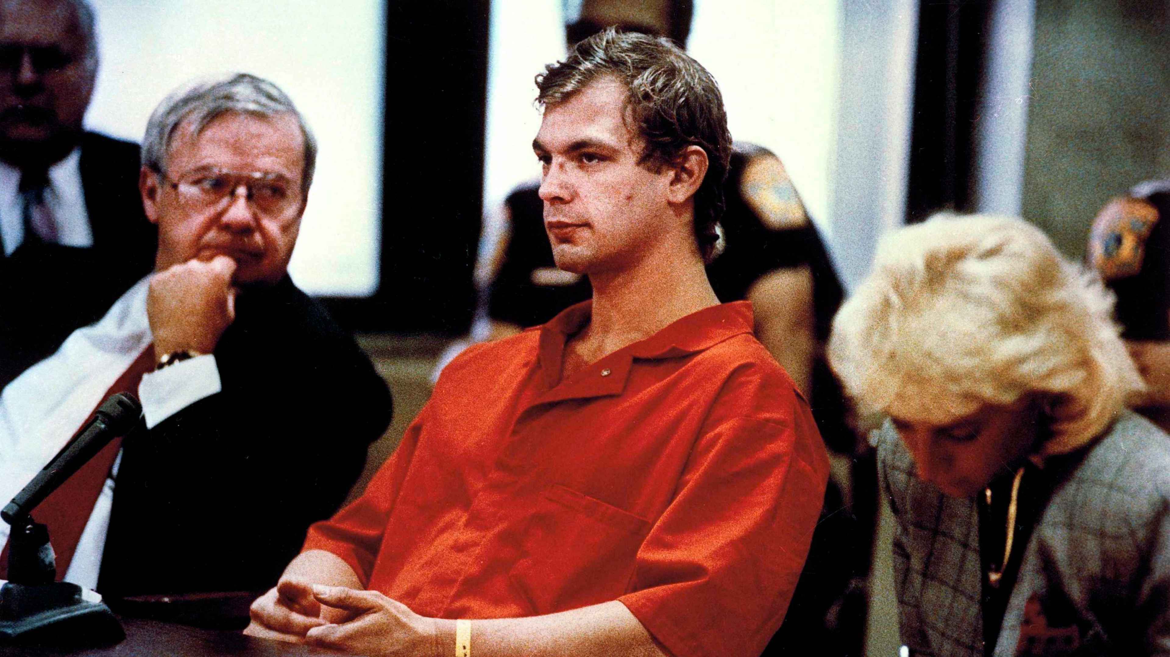 The image shows a man in an orange jumpsuit standing in a courtroom, with two other individuals, likely lawyers or officials, standing behind him.