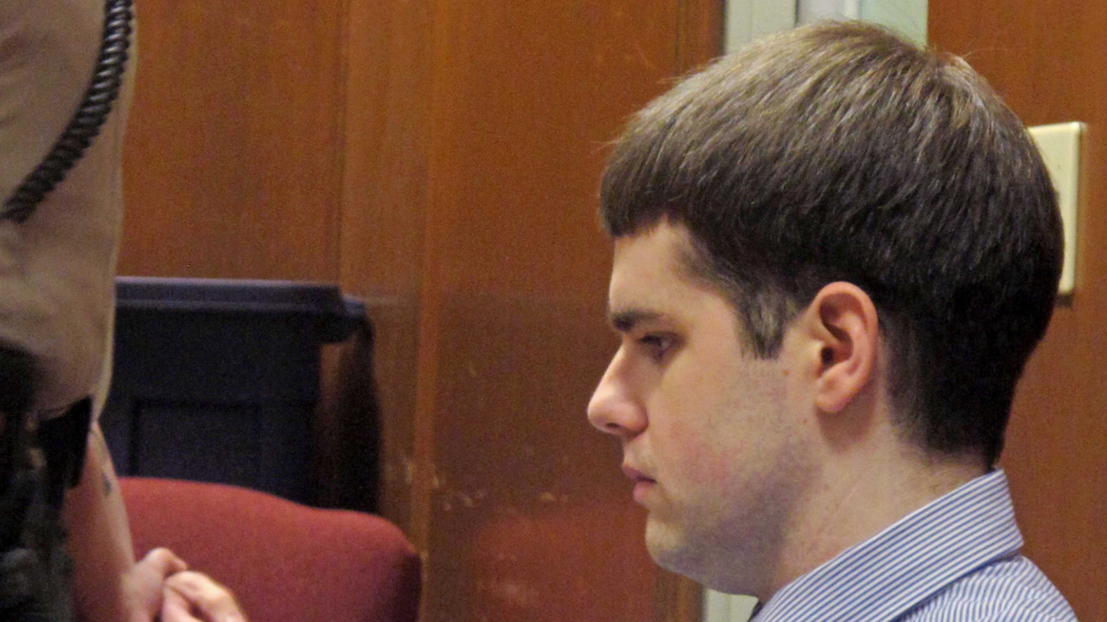 A young man in a suit and tie is sitting in a courtroom, with a wooden bench and wall visible in the background.
