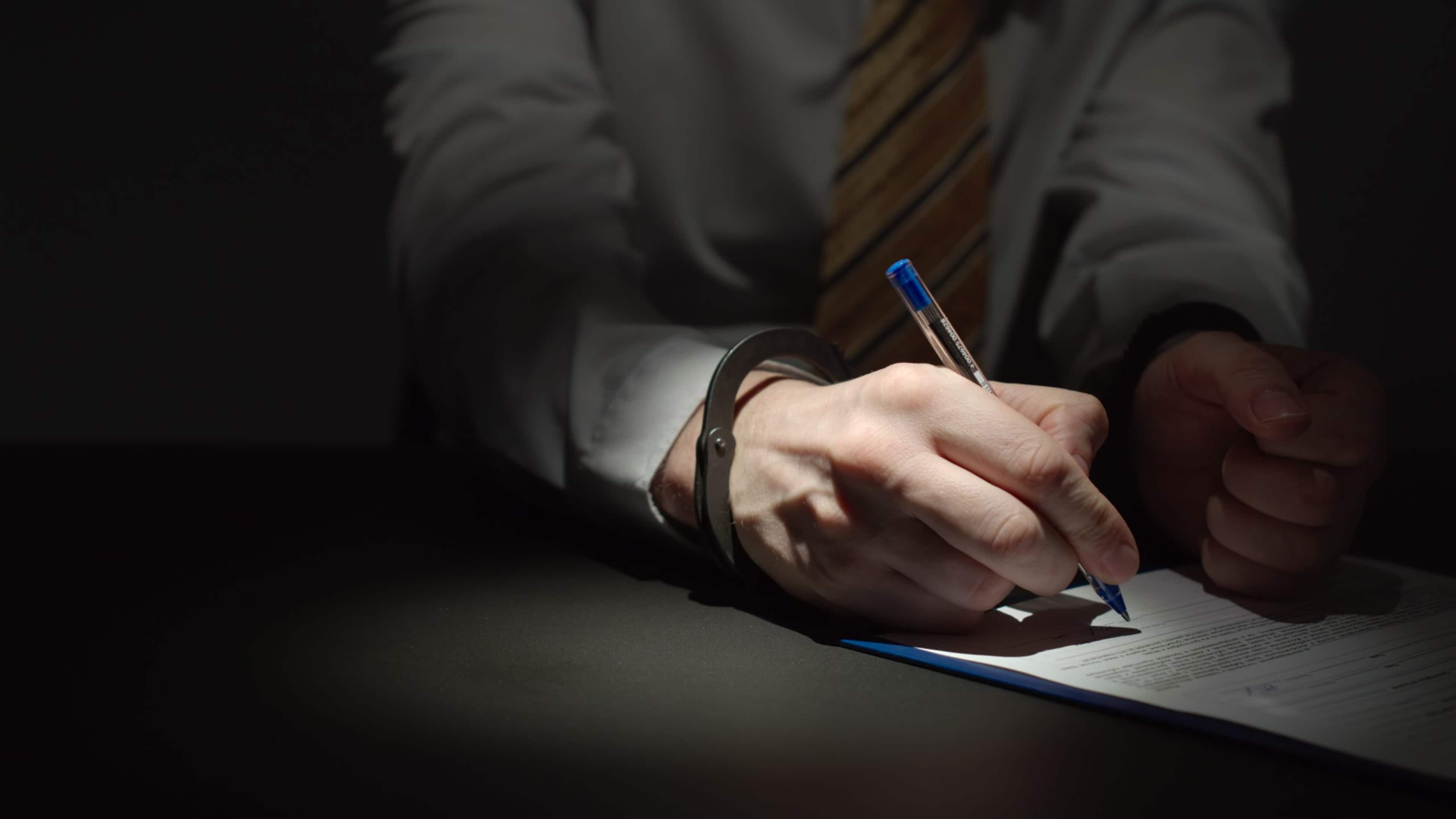 A person's hands holding a pencil and writing on a document in a dimly lit environment.