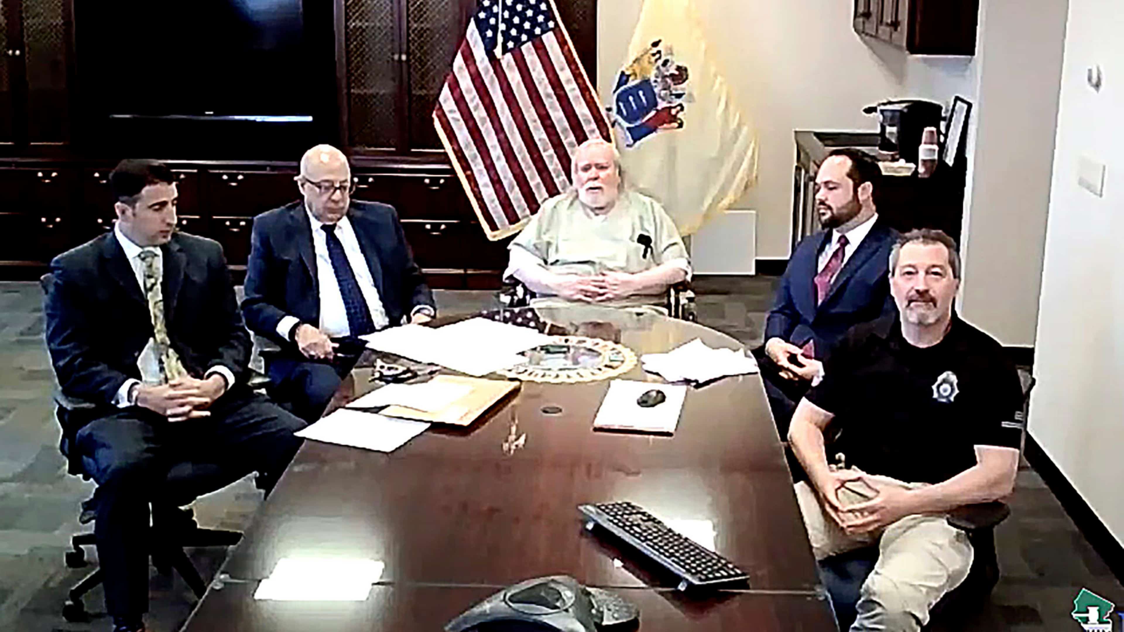 A group of men, some in suits and others in casual attire, are gathered around a table in what appears to be an office or government setting, with an American flag visible in the background.