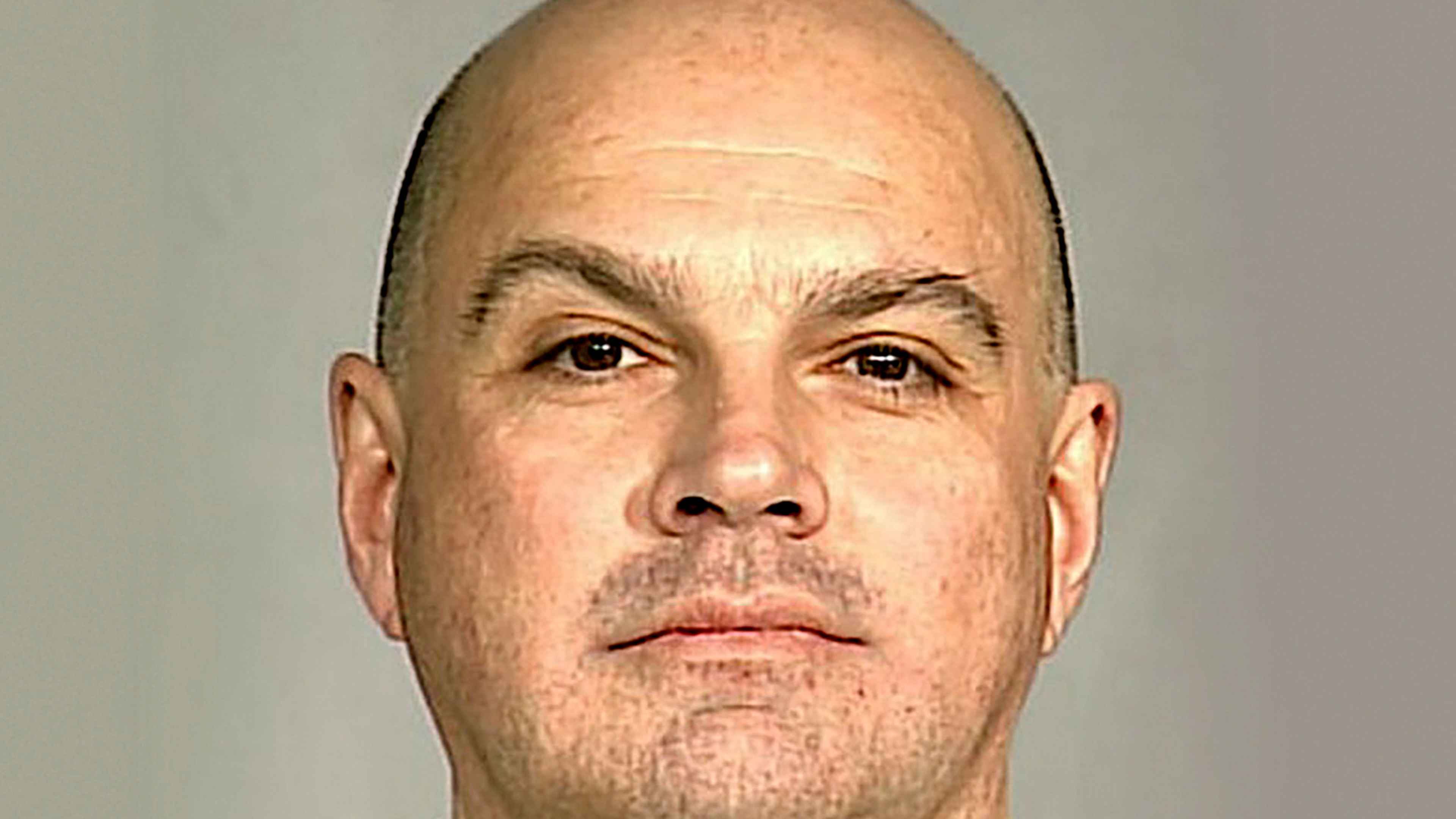 The image shows a close-up portrait of a bald, middle-aged man with a serious expression on his face, set against a plain, light-colored background.