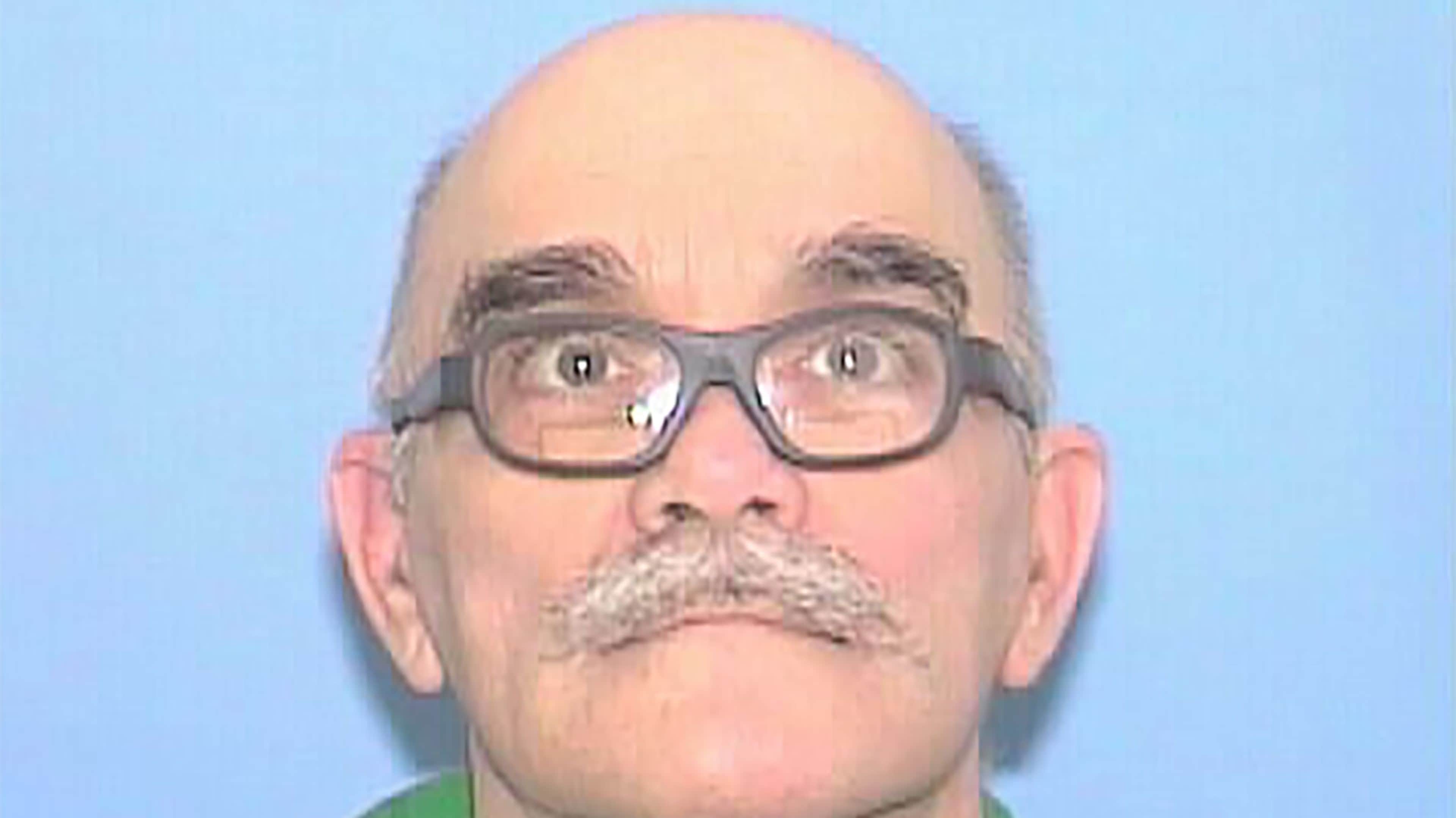 A close-up portrait of a middle-aged man with a mustache and glasses against a plain blue background.