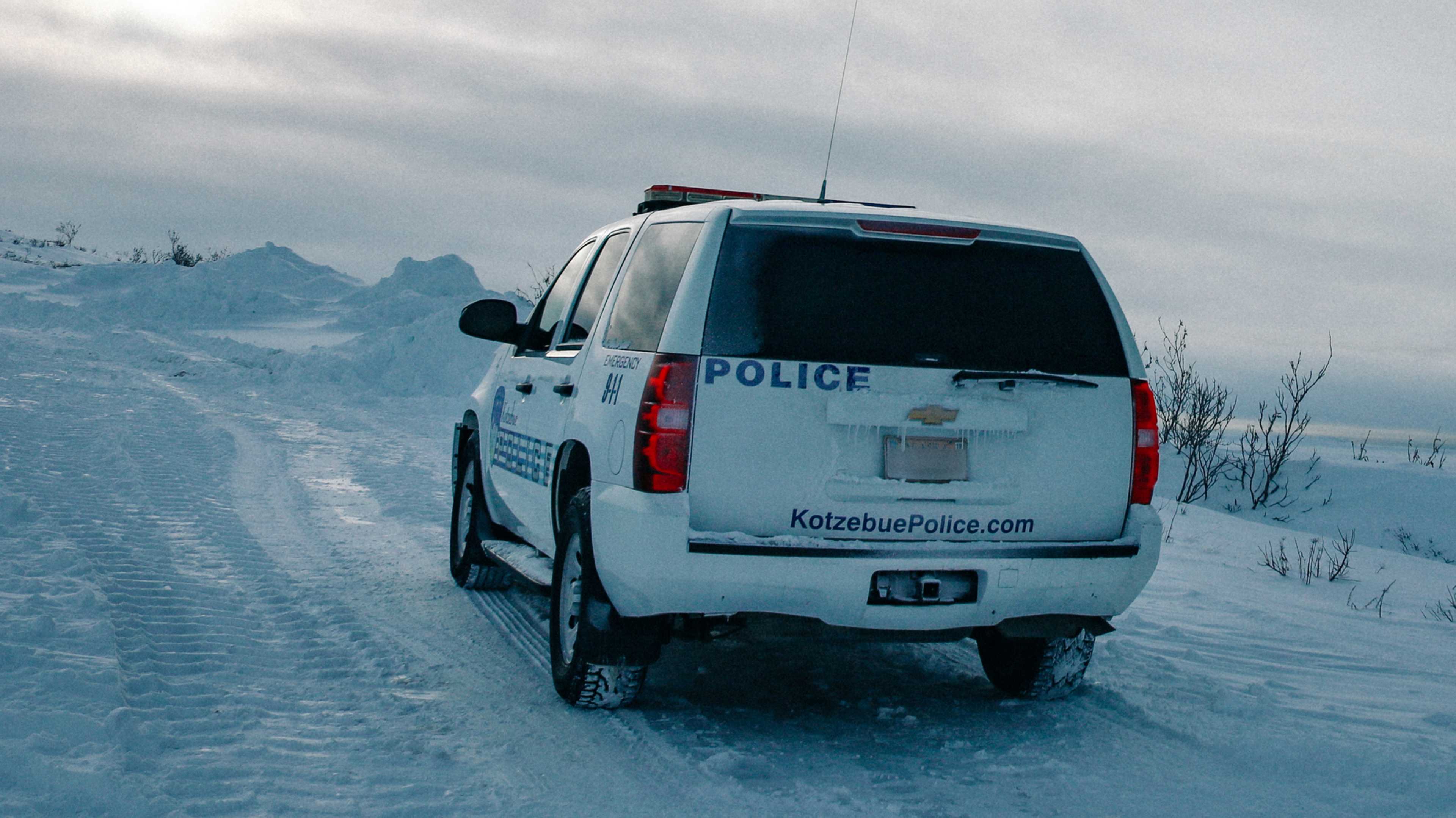 A white police vehicle is parked on a snowy road, with mountains visible in the background.