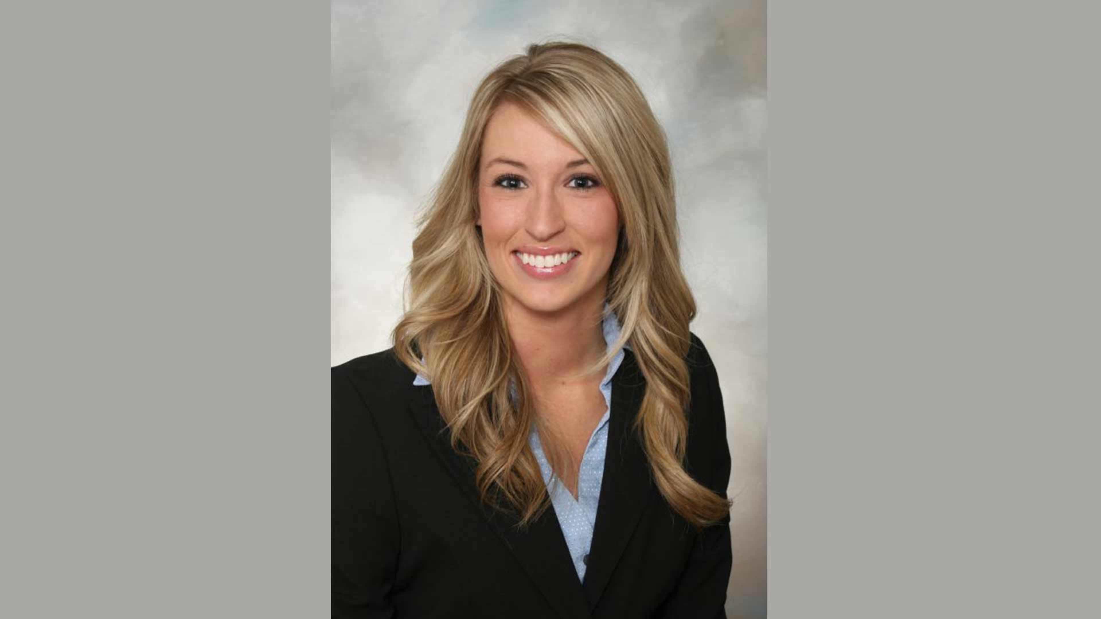 A smiling woman with long, wavy blonde hair wearing a black blazer against a plain background.