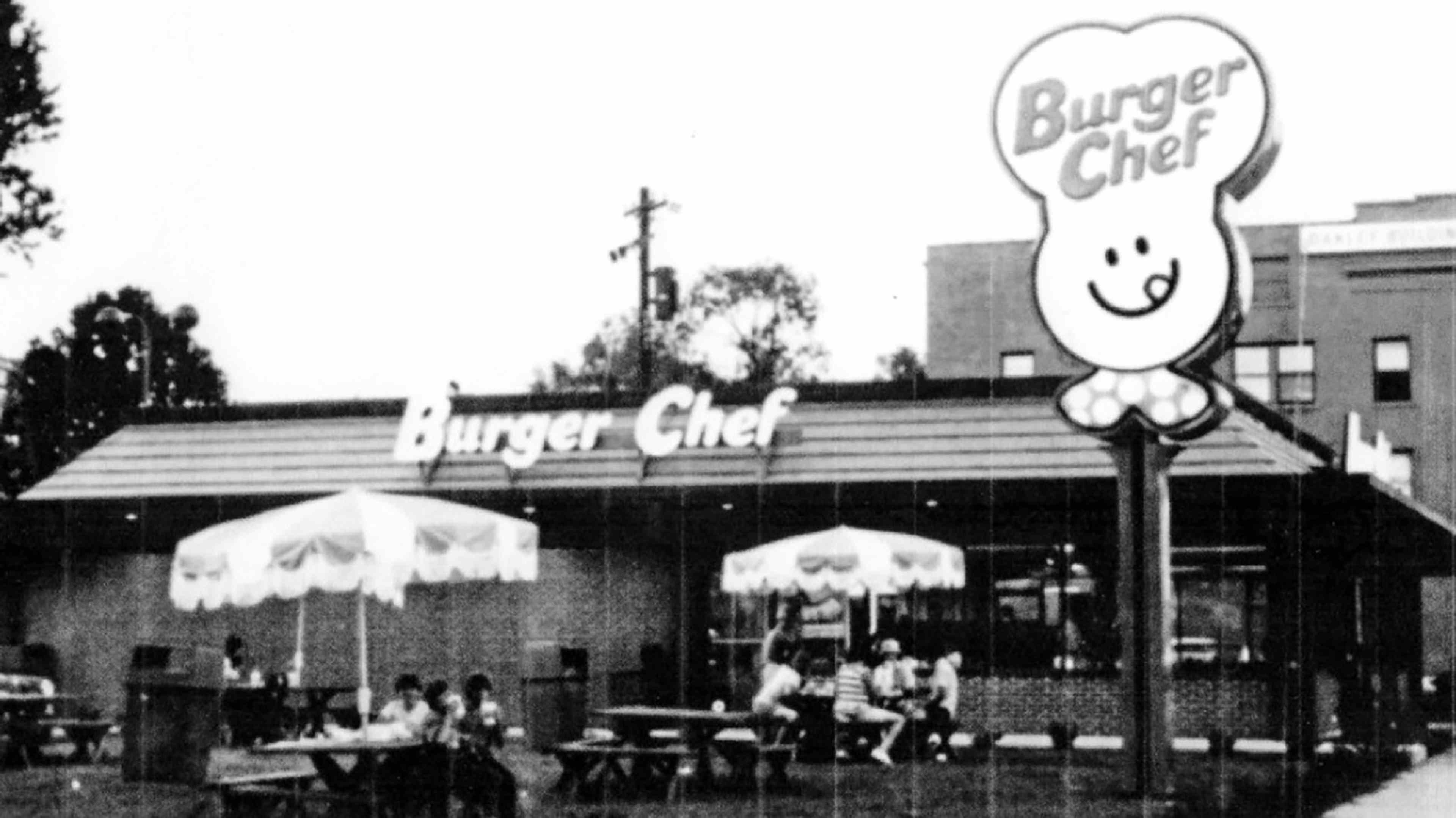 A black and white image of a Burger Chef restaurant, with its iconic smiling chef logo prominently displayed on the building's exterior, surrounded by trees and other structures in the background.
