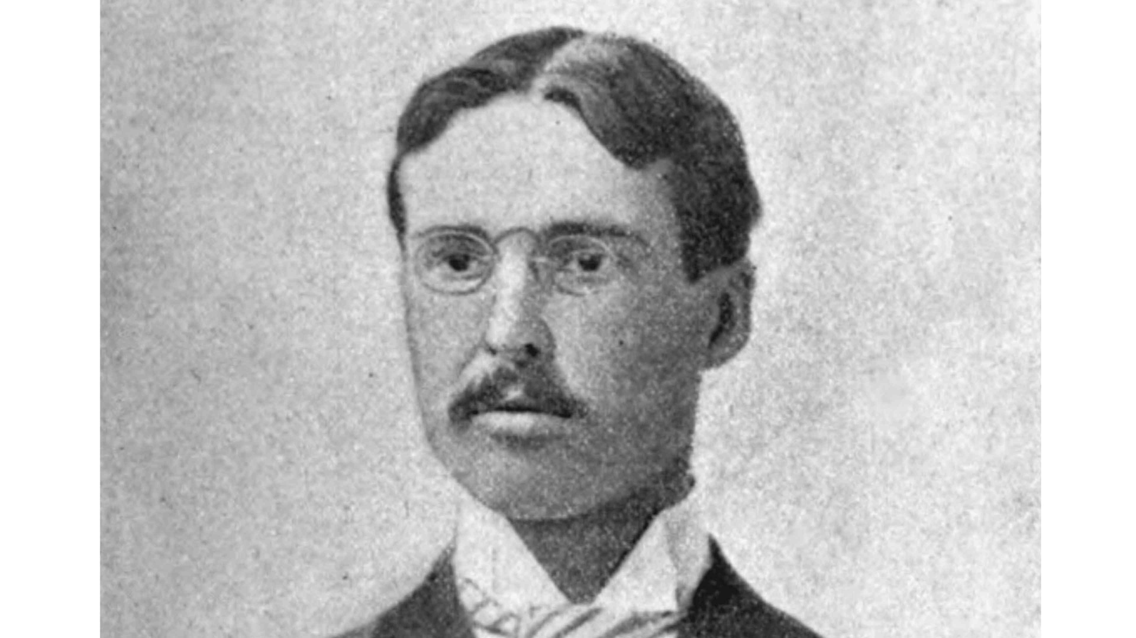 A black and white portrait of a man with a serious expression, wearing a suit and tie, against a plain background.