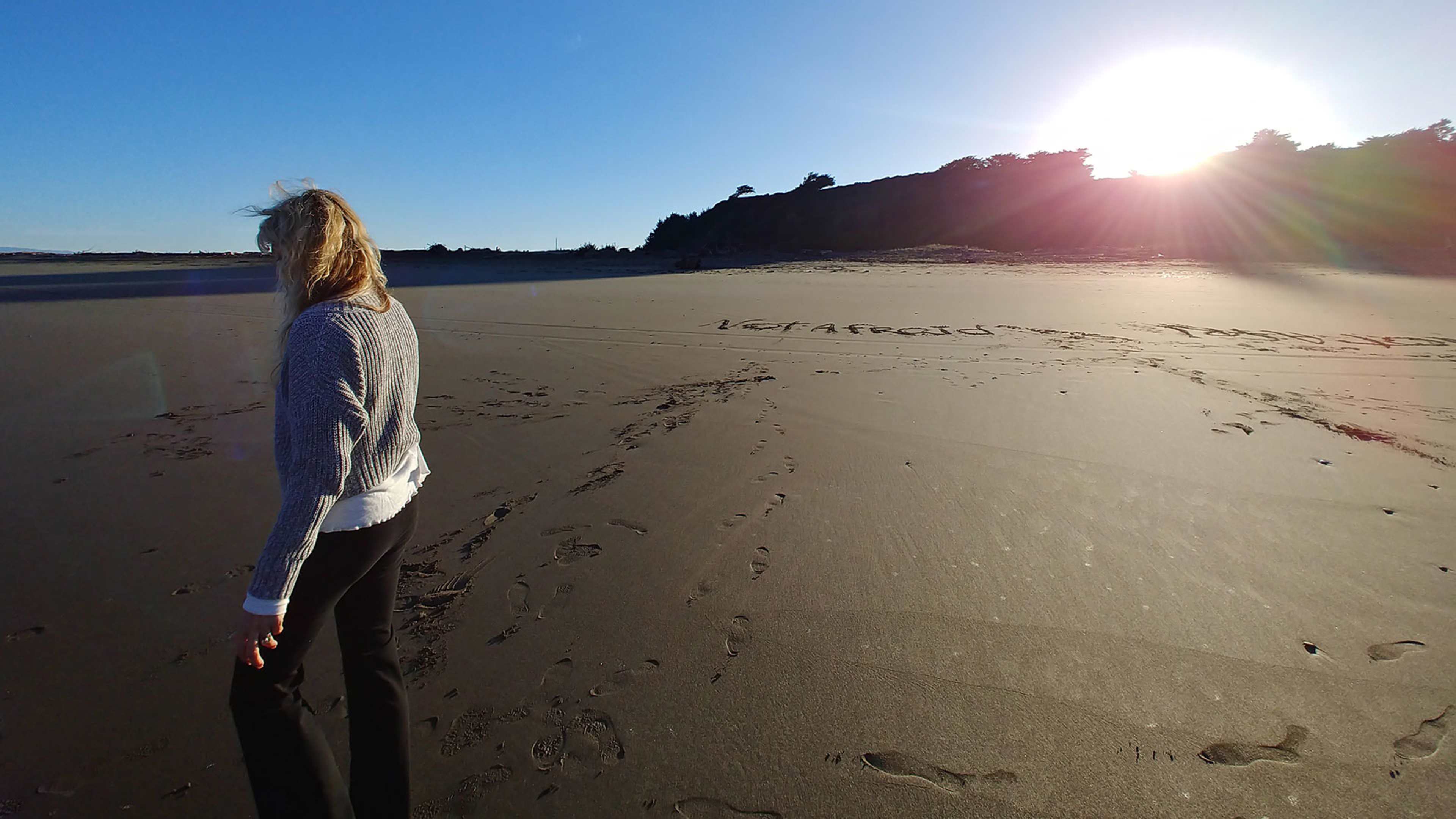 A person with long, blonde hair stands on a sandy beach, facing the ocean as the sun sets behind the rocky cliffs in the distance.