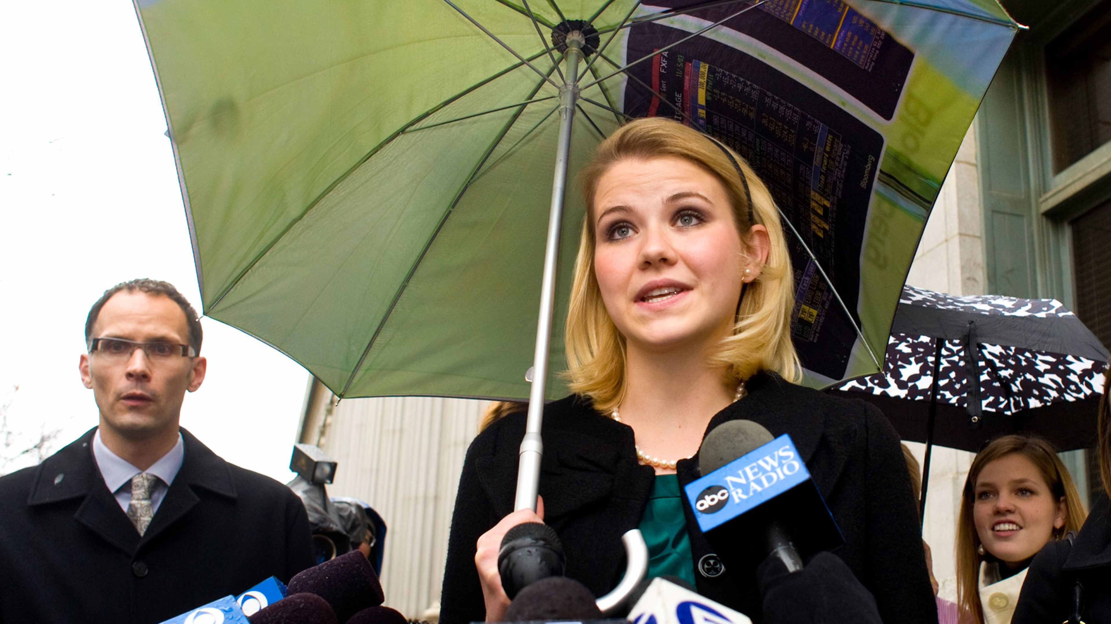 The image shows a woman with a microphone standing in front of a group of people, holding an umbrella and appearing to be giving a news report or interview.