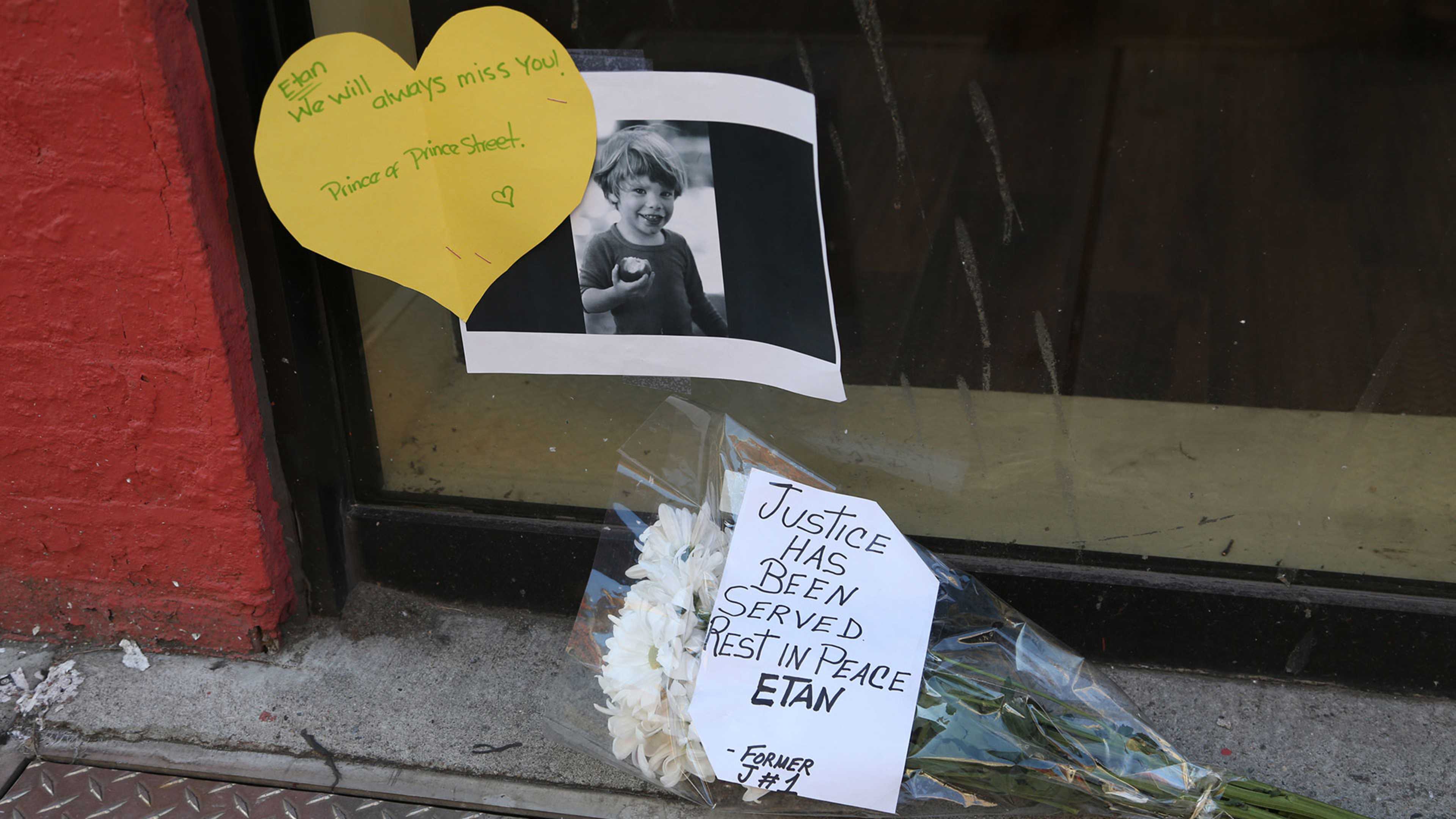 A yellow heart-shaped note with a handwritten message is displayed in the foreground, and a framed photograph of a person is visible in the background. Flowers and a sign with a message are also present in the image.