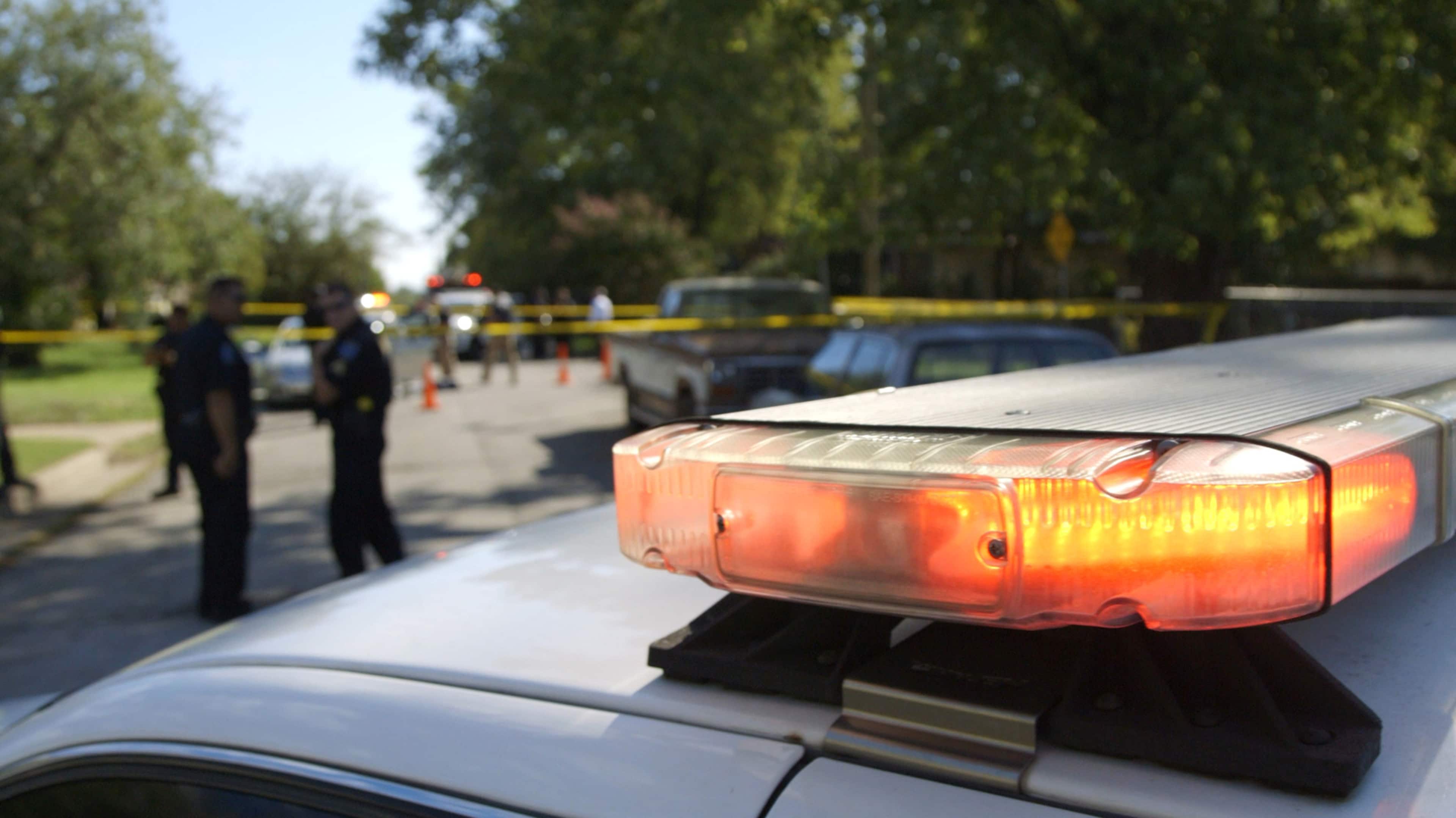 The image shows a police car with its emergency lights flashing, parked on a street surrounded by trees and people in the background.