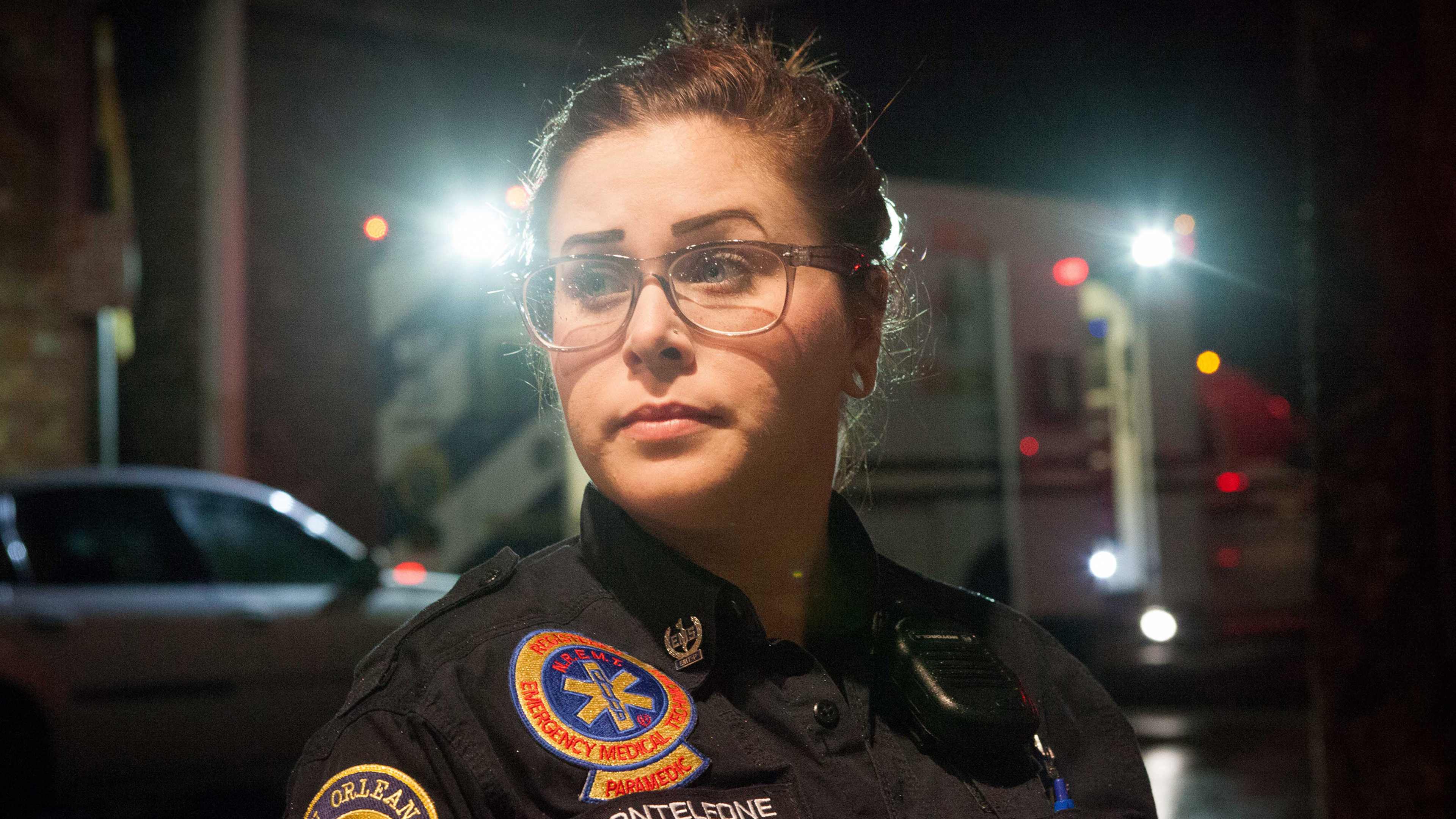 A female police officer wearing a uniform and glasses stands in a dimly lit urban setting, with the lights of passing vehicles visible in the background.