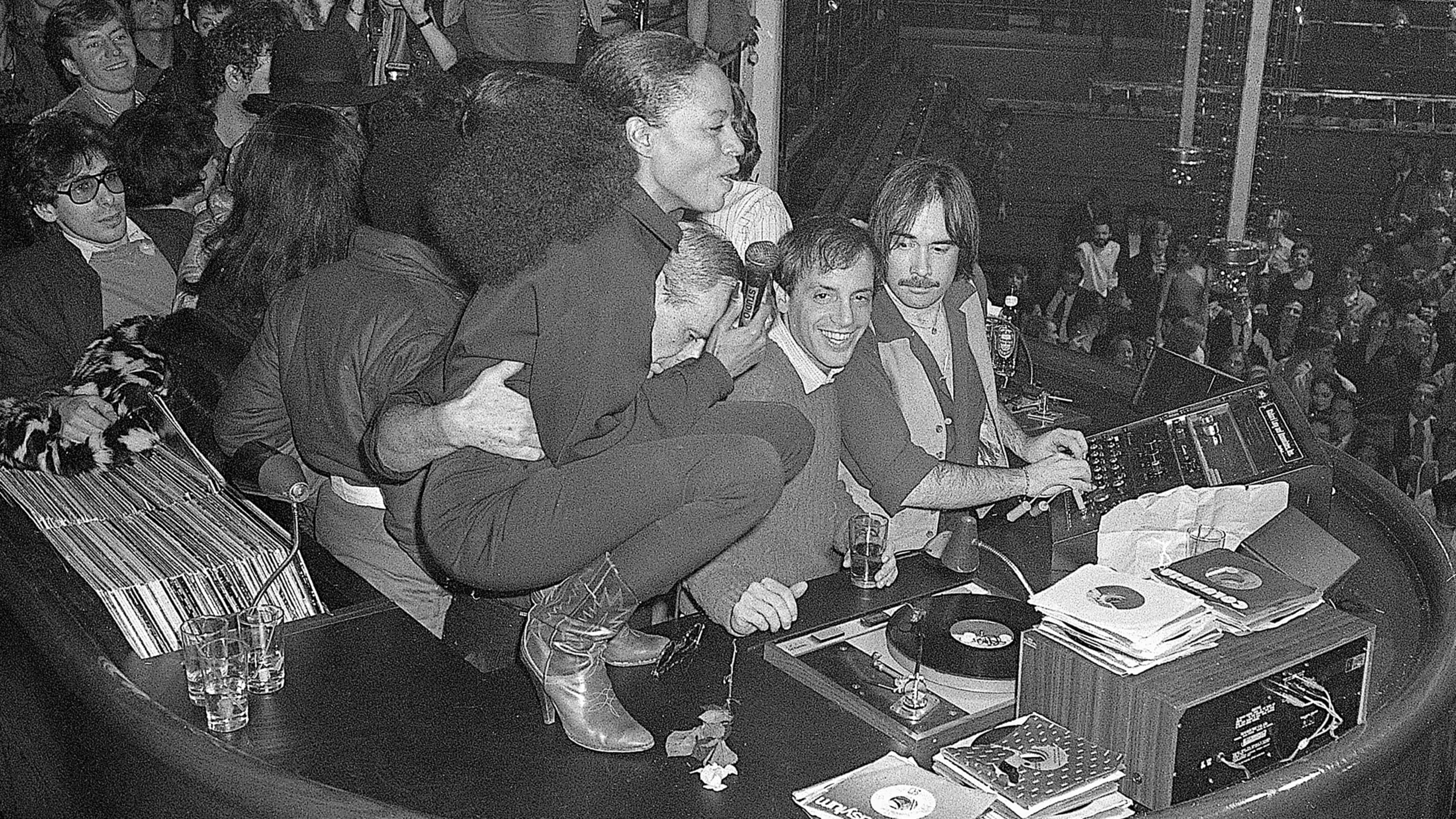 A crowded room filled with people, some sitting on the floor and others standing, surrounded by various musical equipment and records.