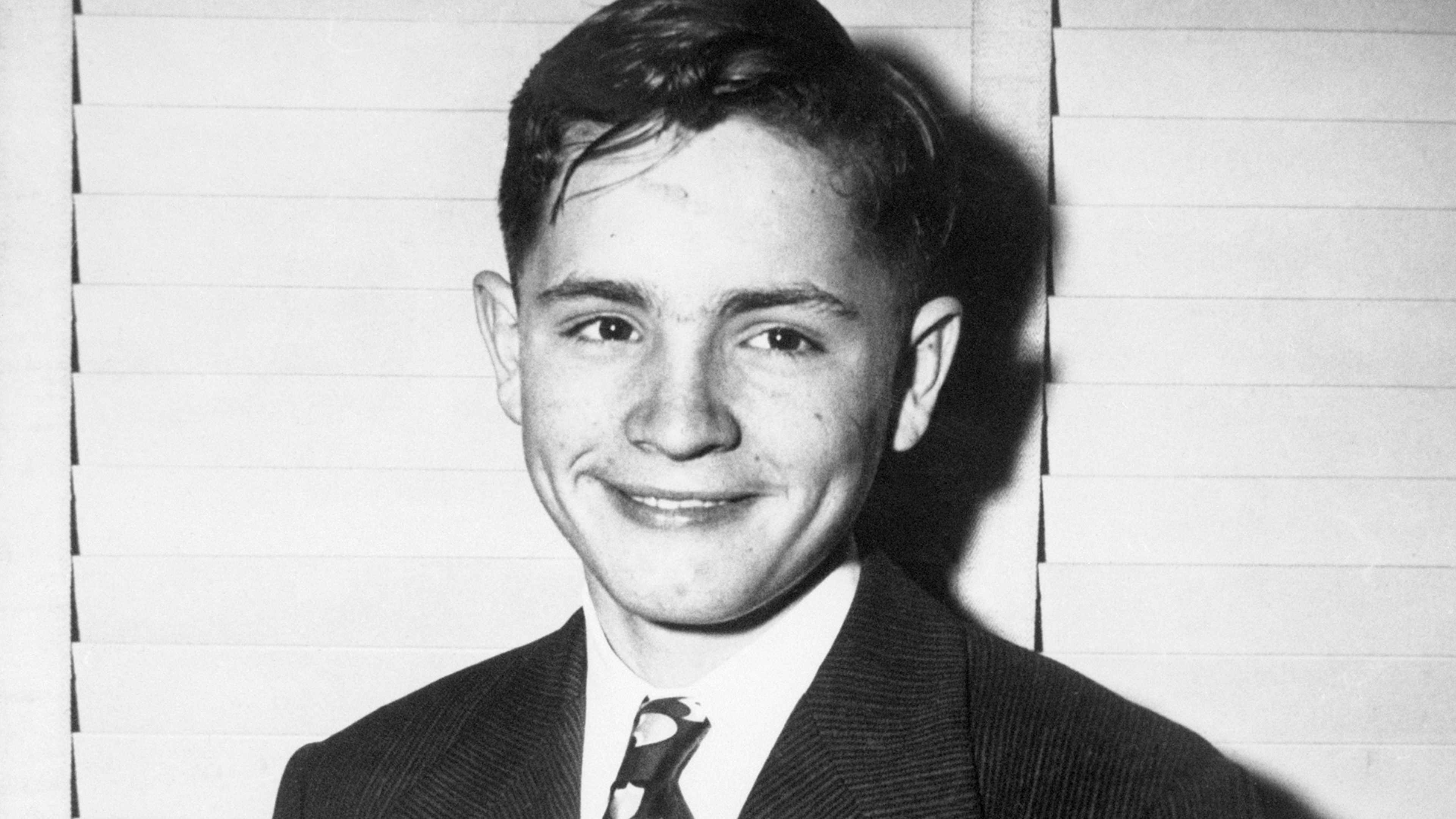 A black and white portrait photograph of a smiling young man with short dark hair, wearing a suit and tie, set against a plain white background.
