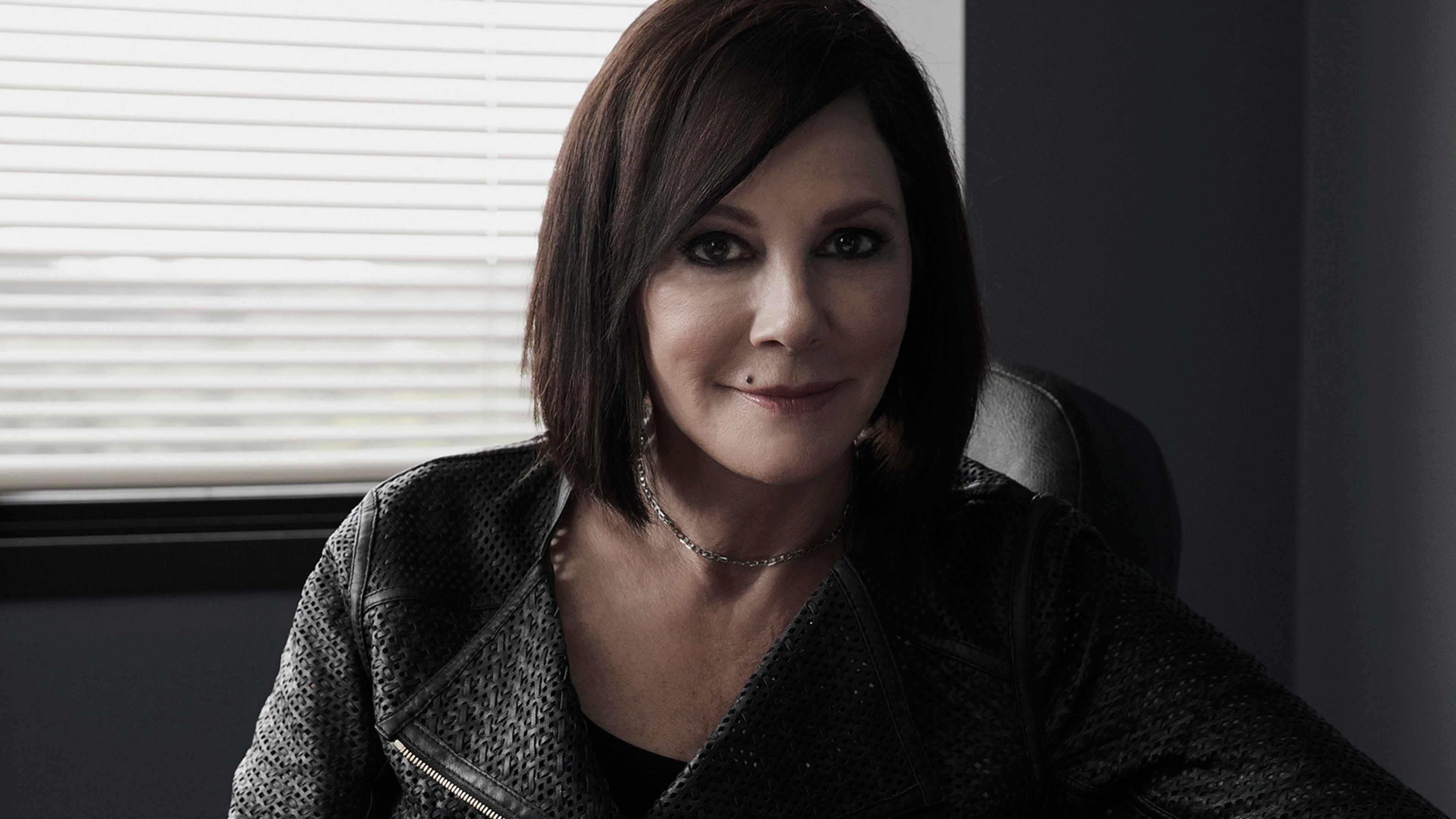 A woman with dark hair and a serious expression is sitting near a window, with a blurred background visible behind her.