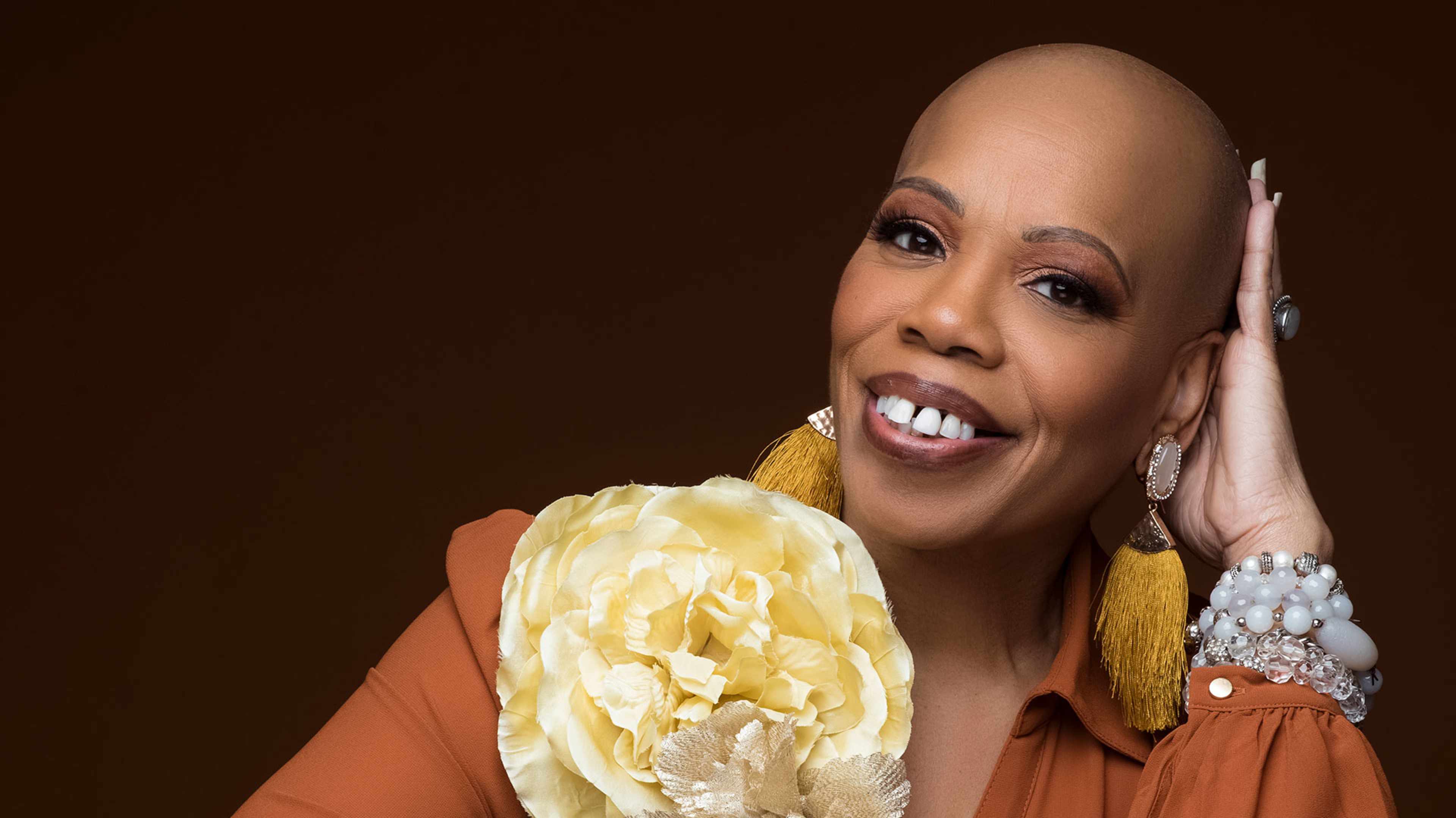 A smiling woman with a vibrant yellow flower in her hand stands against a dark brown background.
