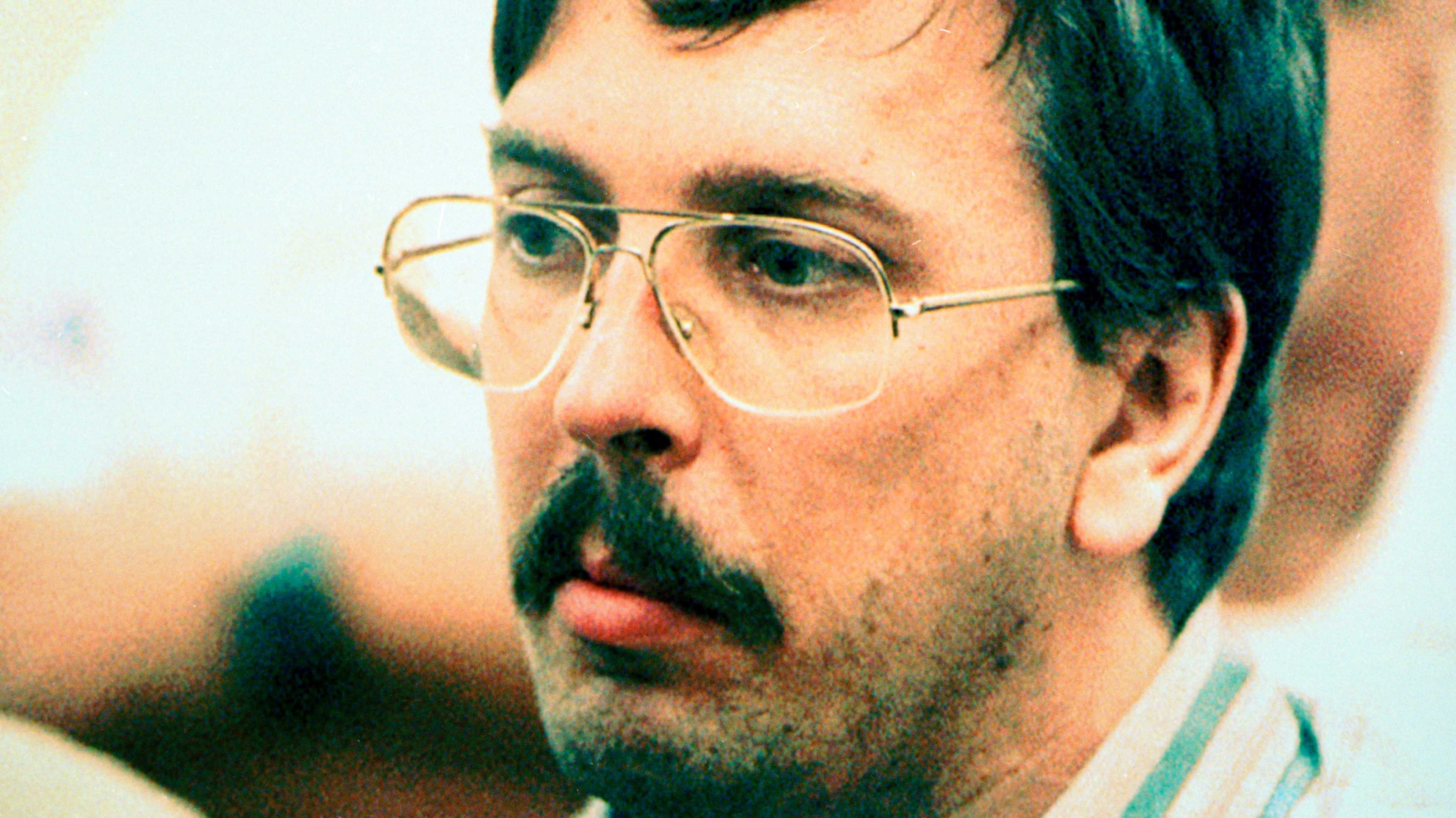 The image shows a close-up portrait of a man with dark hair and glasses, his face appearing thoughtful or pensive against a blurred background.