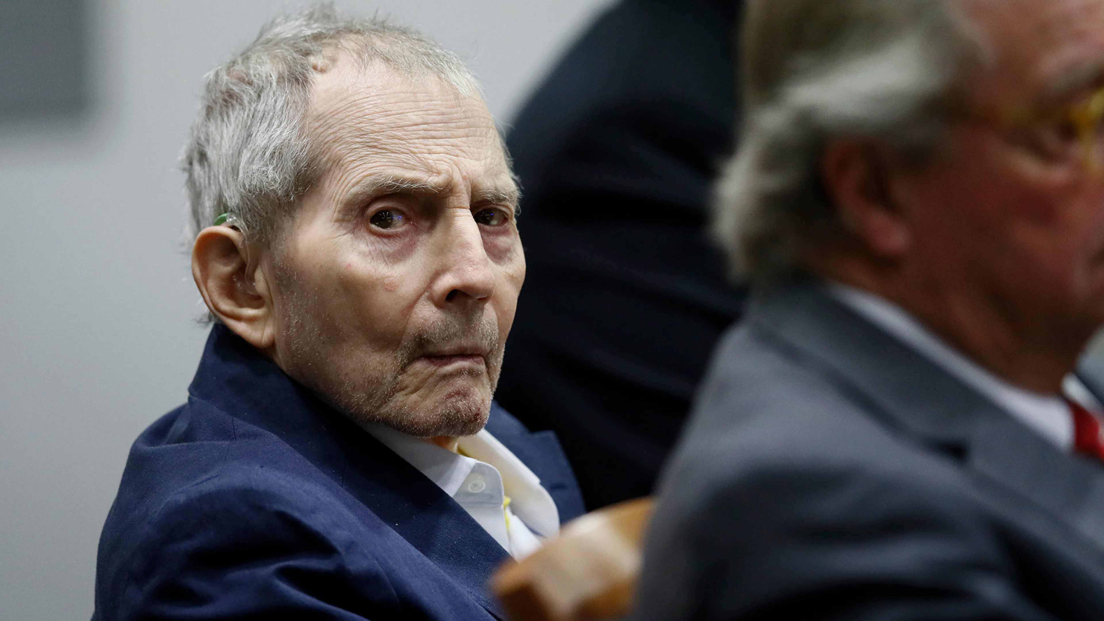 The image shows an elderly man with gray hair and a serious expression, wearing a blue suit and sitting in what appears to be a courtroom or legal setting.