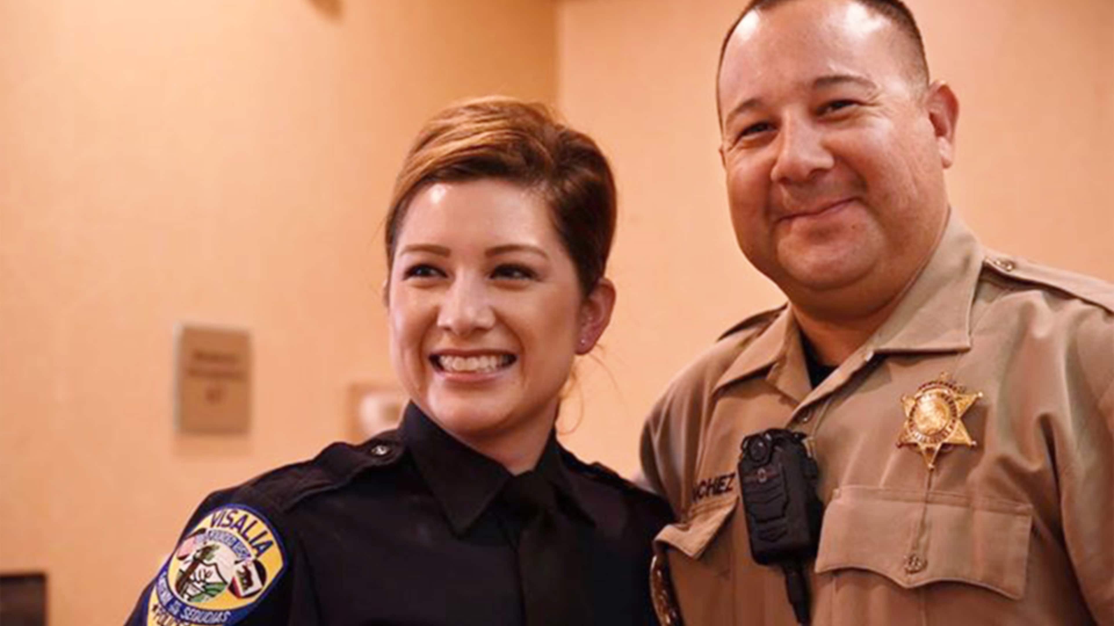 The image shows two law enforcement officers, a woman and a man, standing together and smiling in what appears to be an indoor setting.
