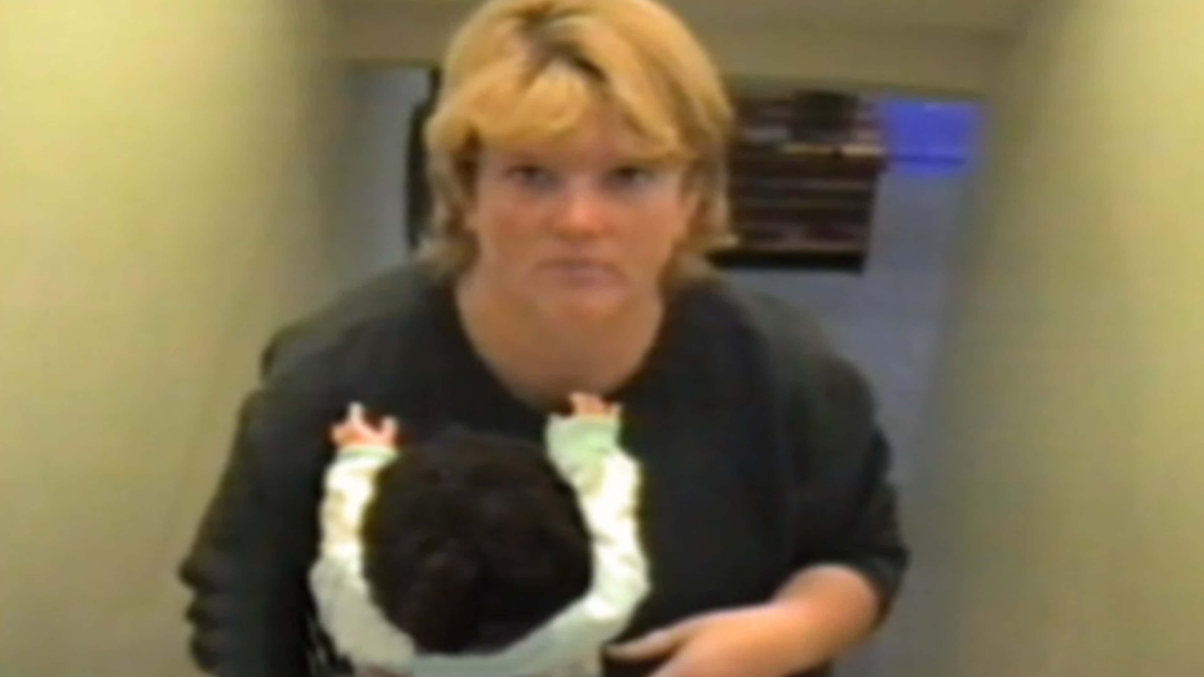 The image shows a woman with blonde hair wearing a black jacket, standing in what appears to be a hallway or indoor setting.