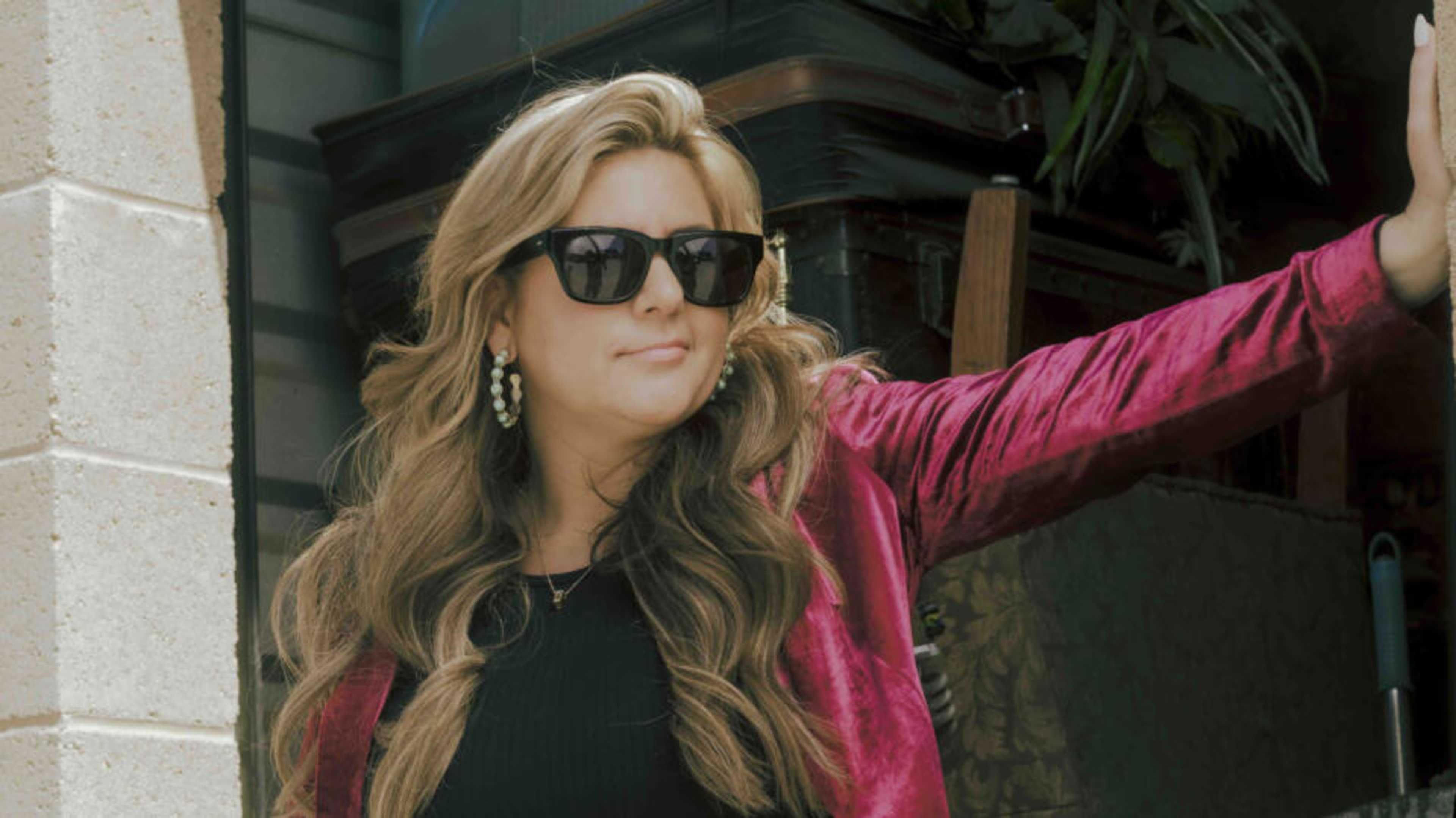 A woman with long, wavy blonde hair wearing sunglasses and a bright pink jacket stands in front of a brick building.