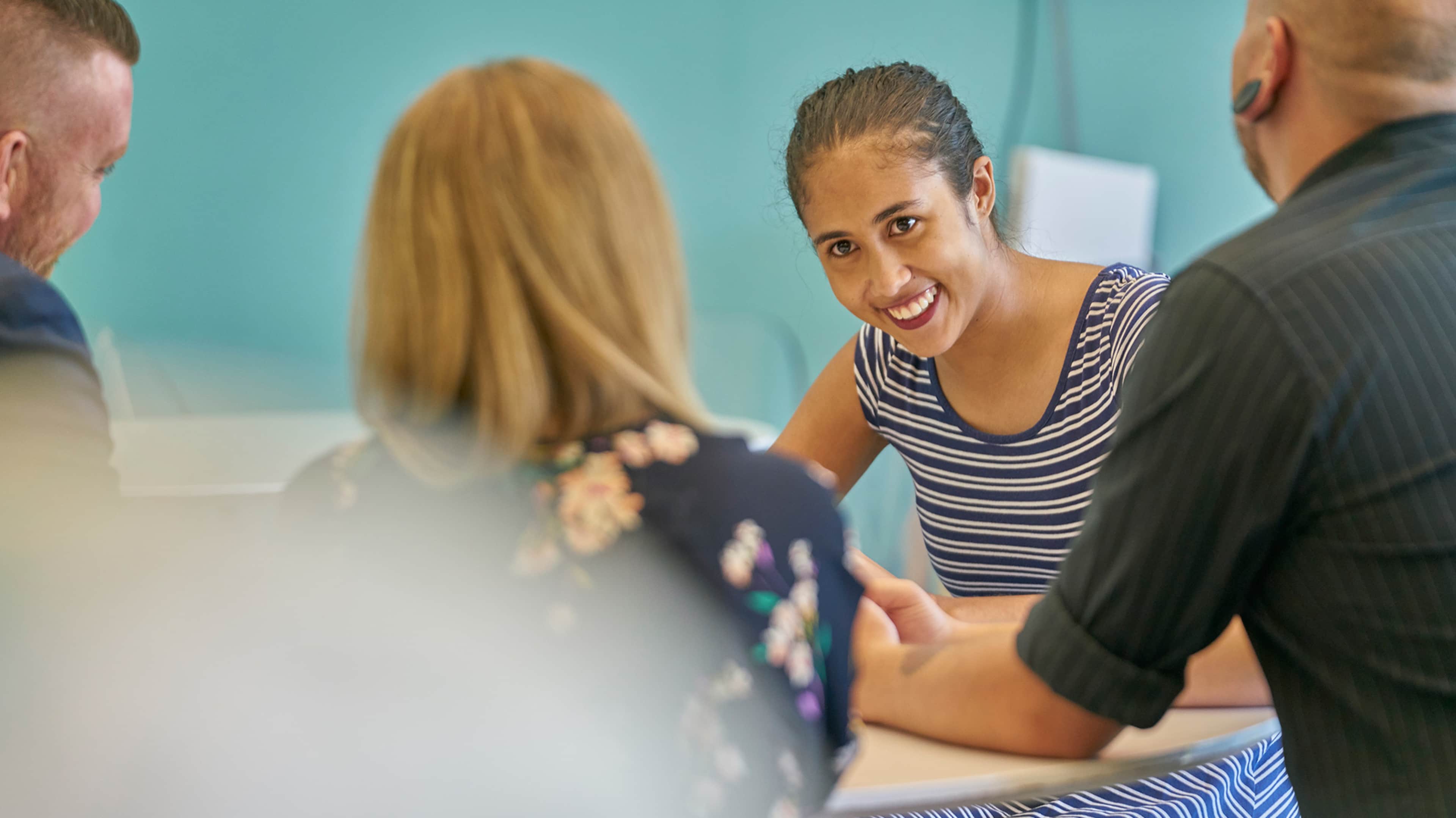 A group of people, including a woman with a striped shirt, engaged in a lively discussion in what appears to be an office or classroom setting.