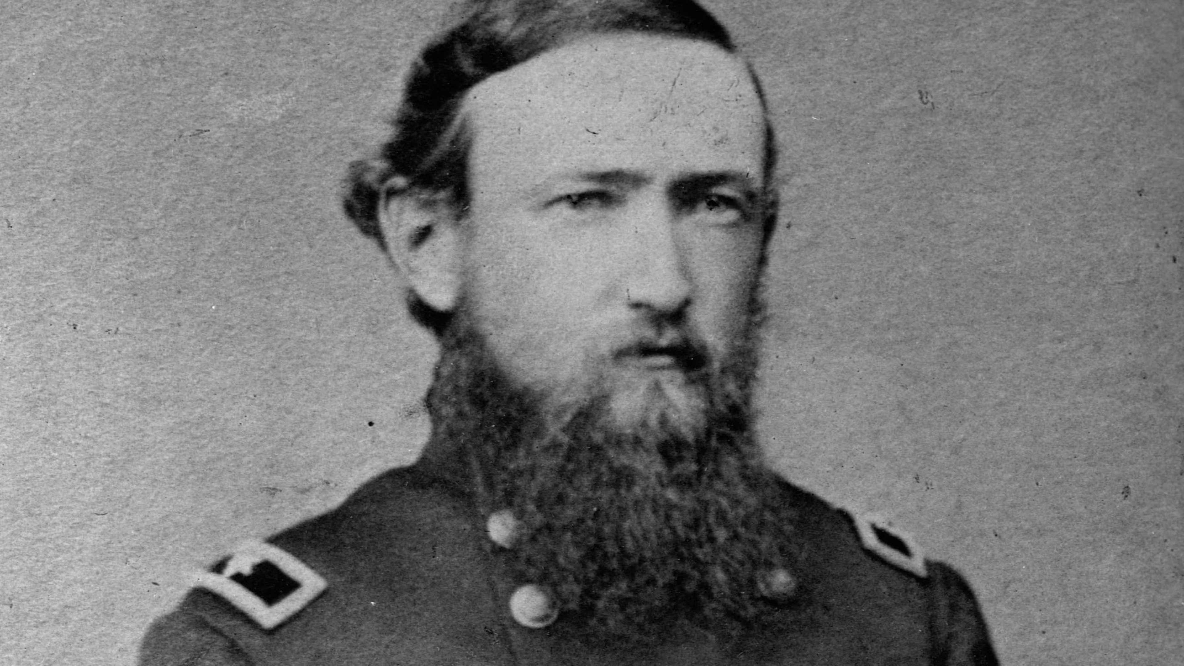 A black and white portrait photograph of a man with a thick beard, wearing a military uniform with epaulets, standing in front of a plain background.