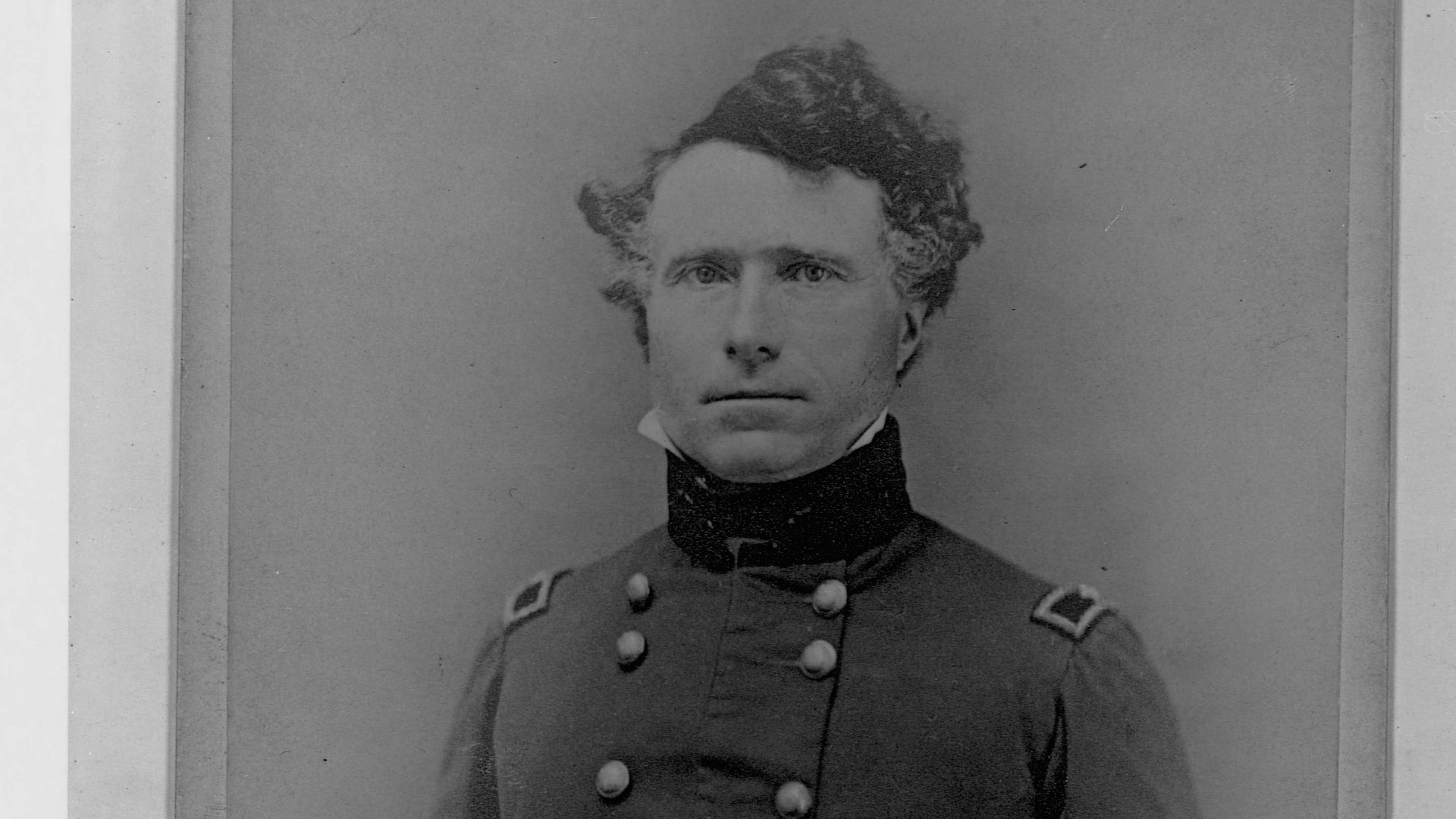 A black and white portrait photograph of a man in military uniform, standing upright with his hands clasped in front of him, against a plain background.