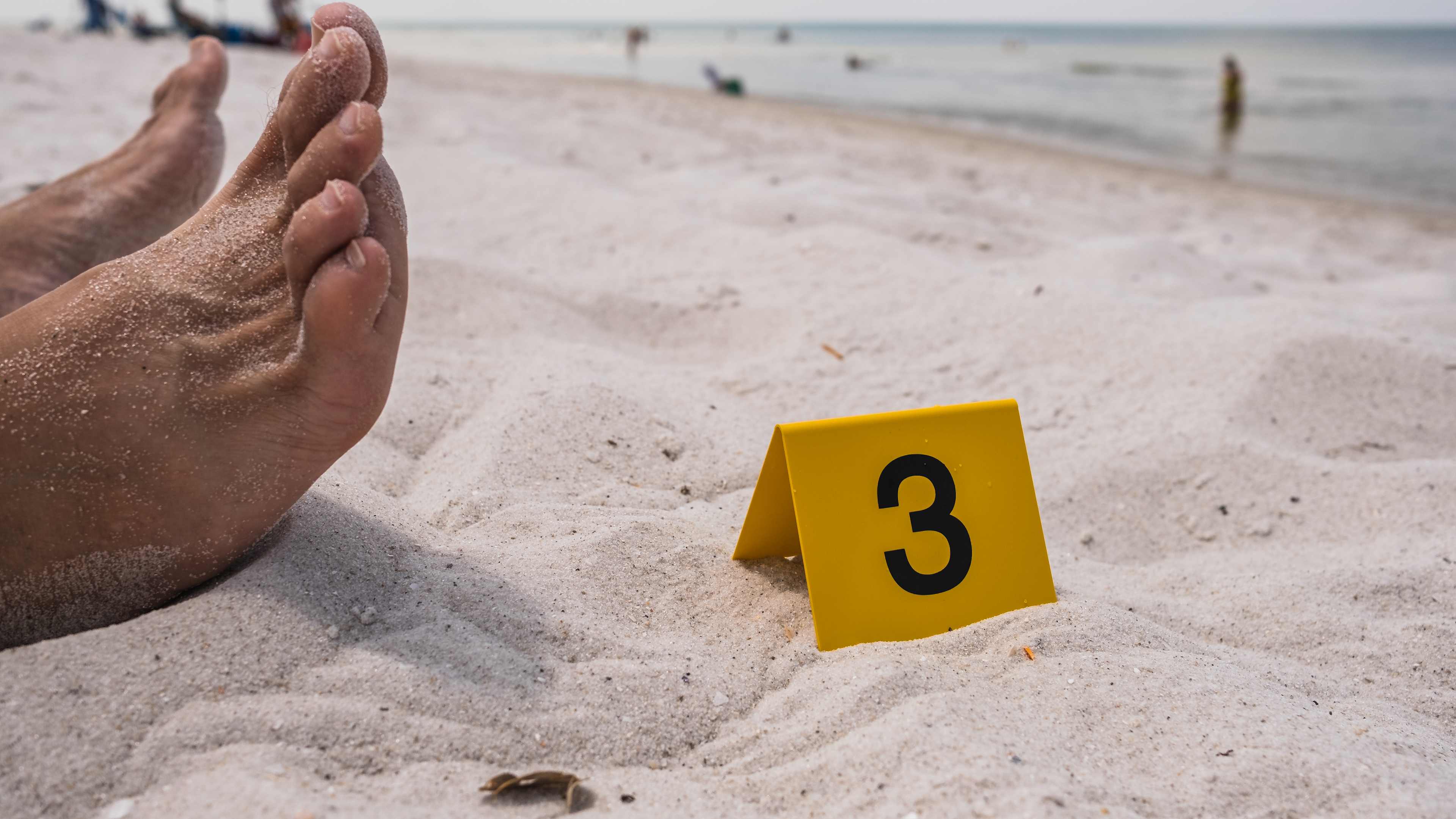 One yellow crime scene evidence marker next to the foot of a corpse laying on the beach after a murder shooting stabbing body forensics csi
