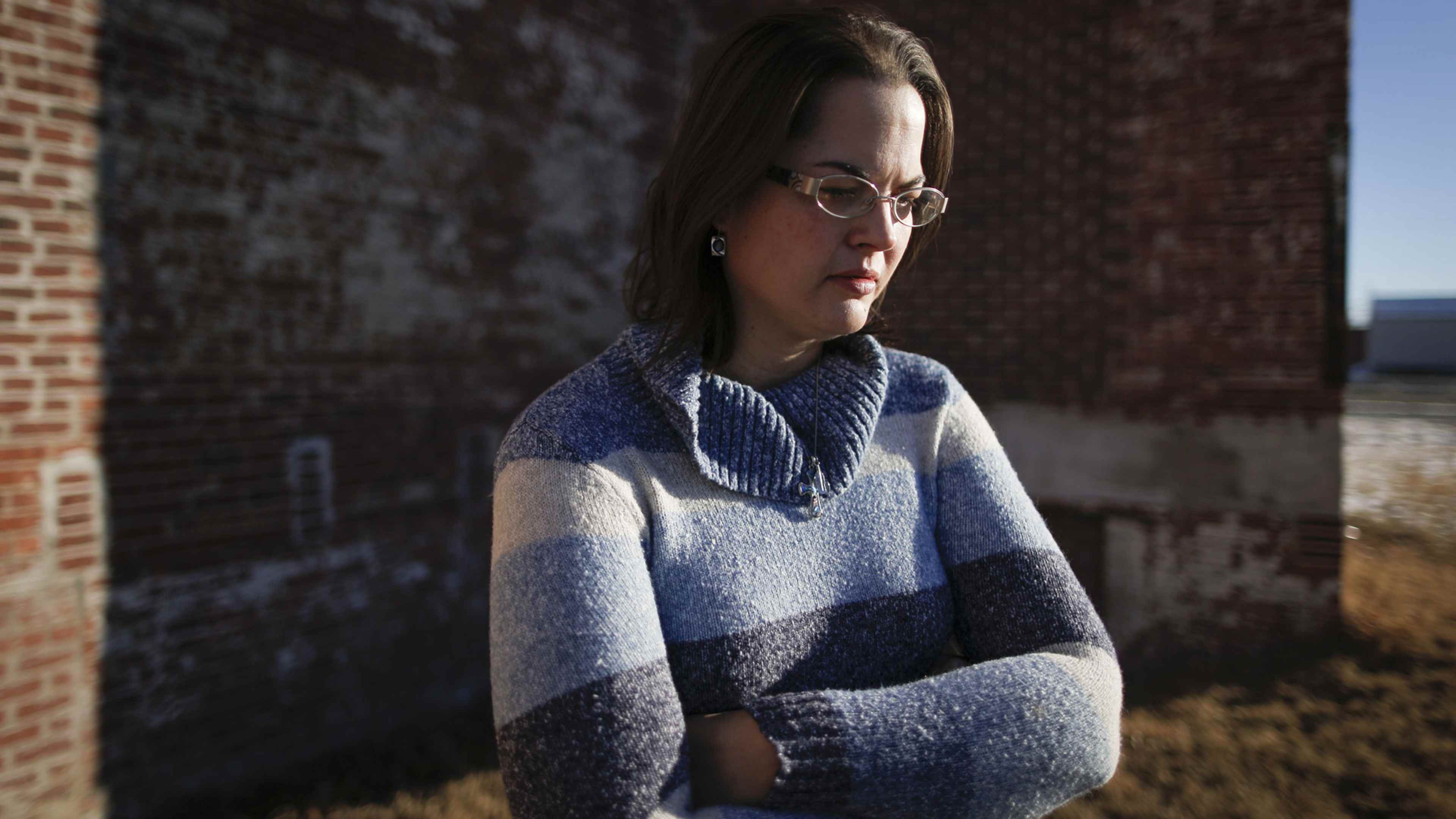 A young woman wearing a blue and gray sweater stands in front of a brick wall, her expression thoughtful as she gazes off to the side.