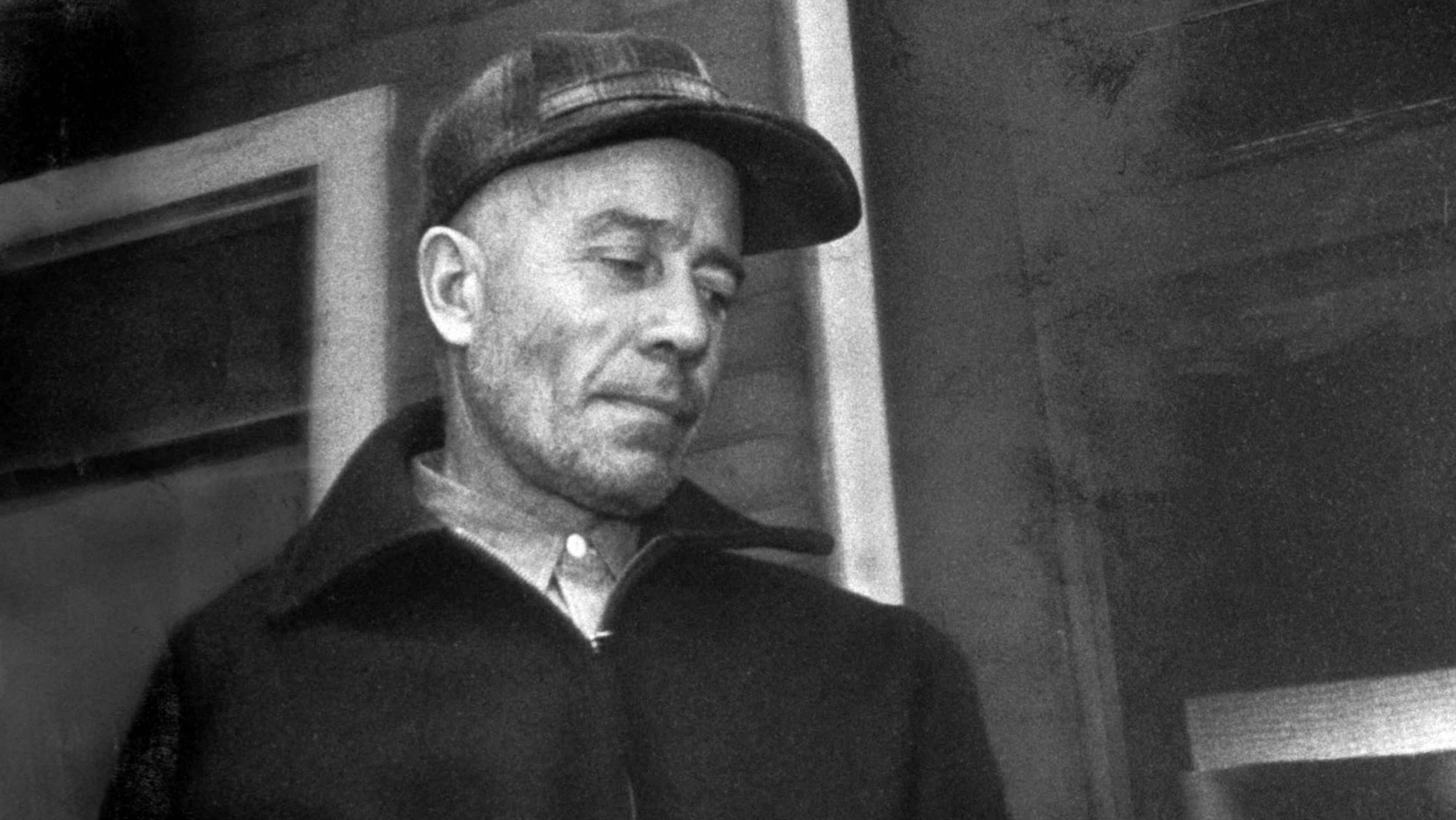 A weathered, elderly man with a cap stands in a dimly lit doorway, his face etched with lines and his expression somber.