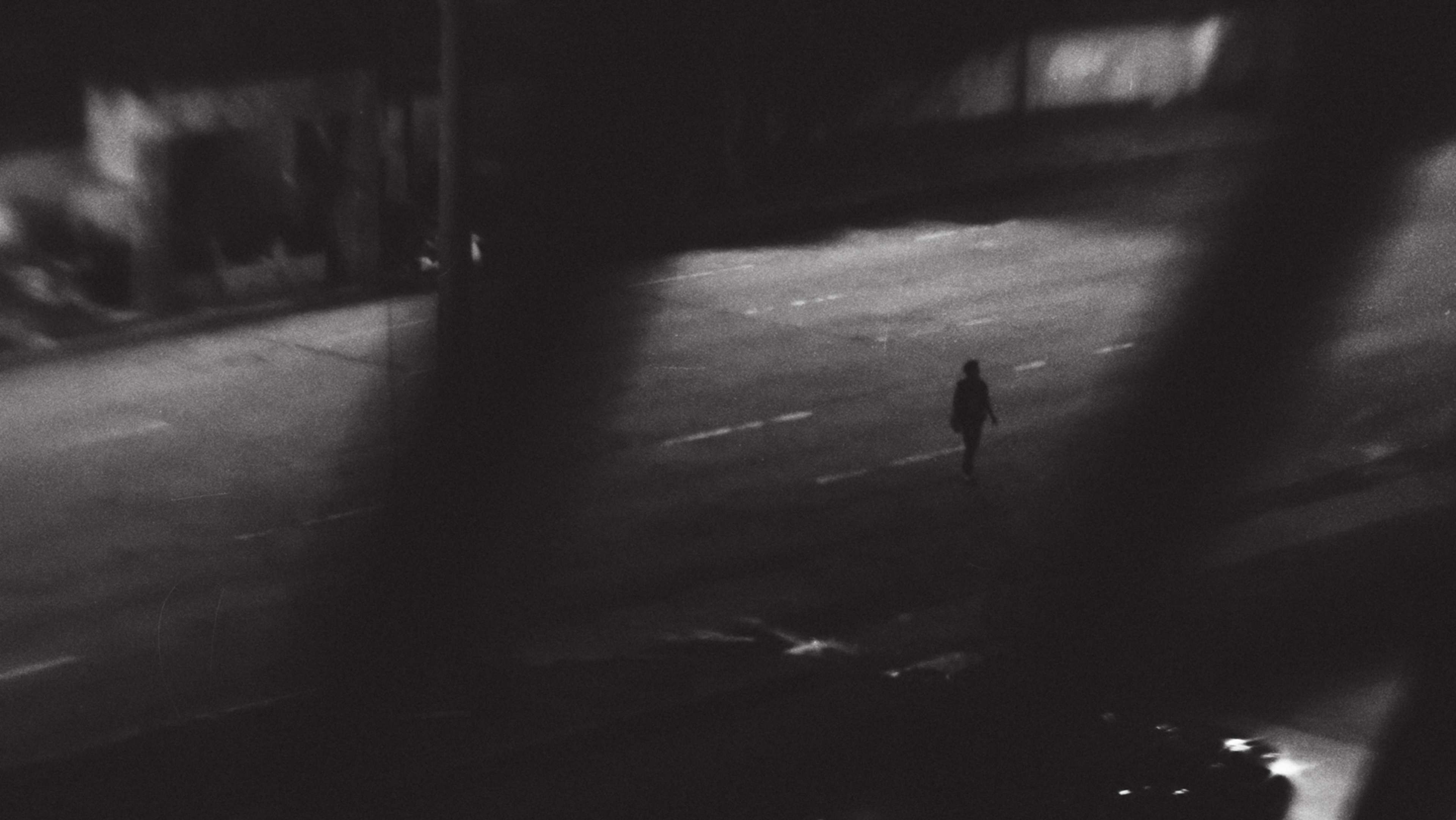A solitary figure walks down a dimly lit, shadowy alley, the surroundings obscured in darkness.