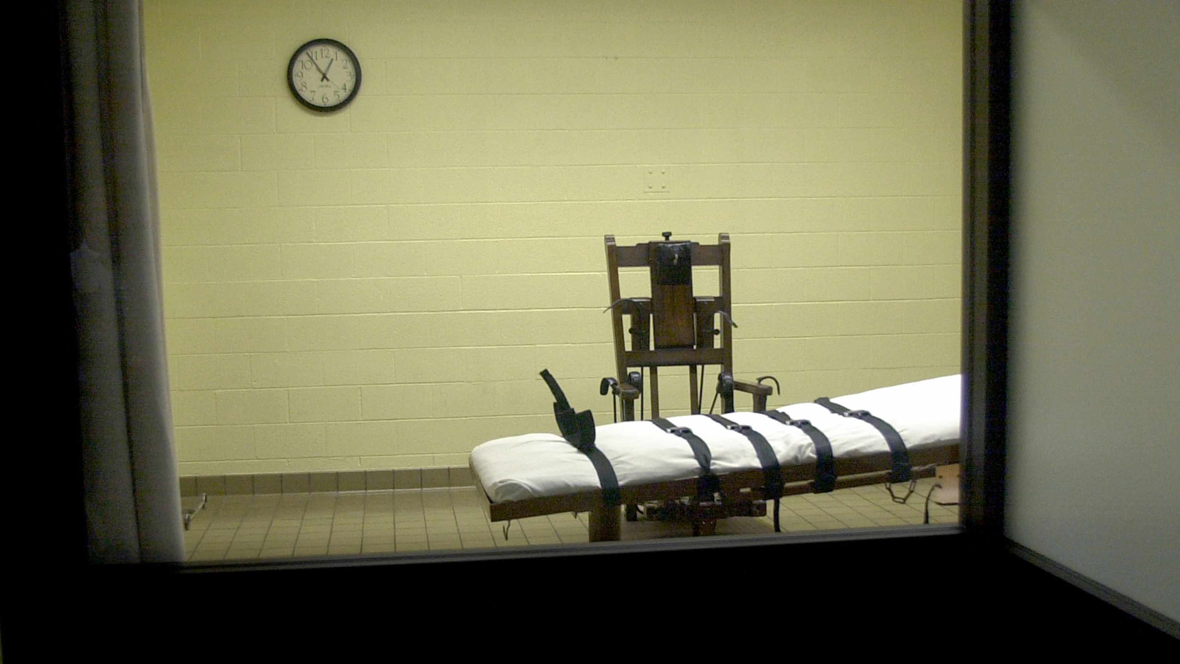 A hospital bed with medical equipment stands in a dimly lit room, with a clock visible on the wall in the background.