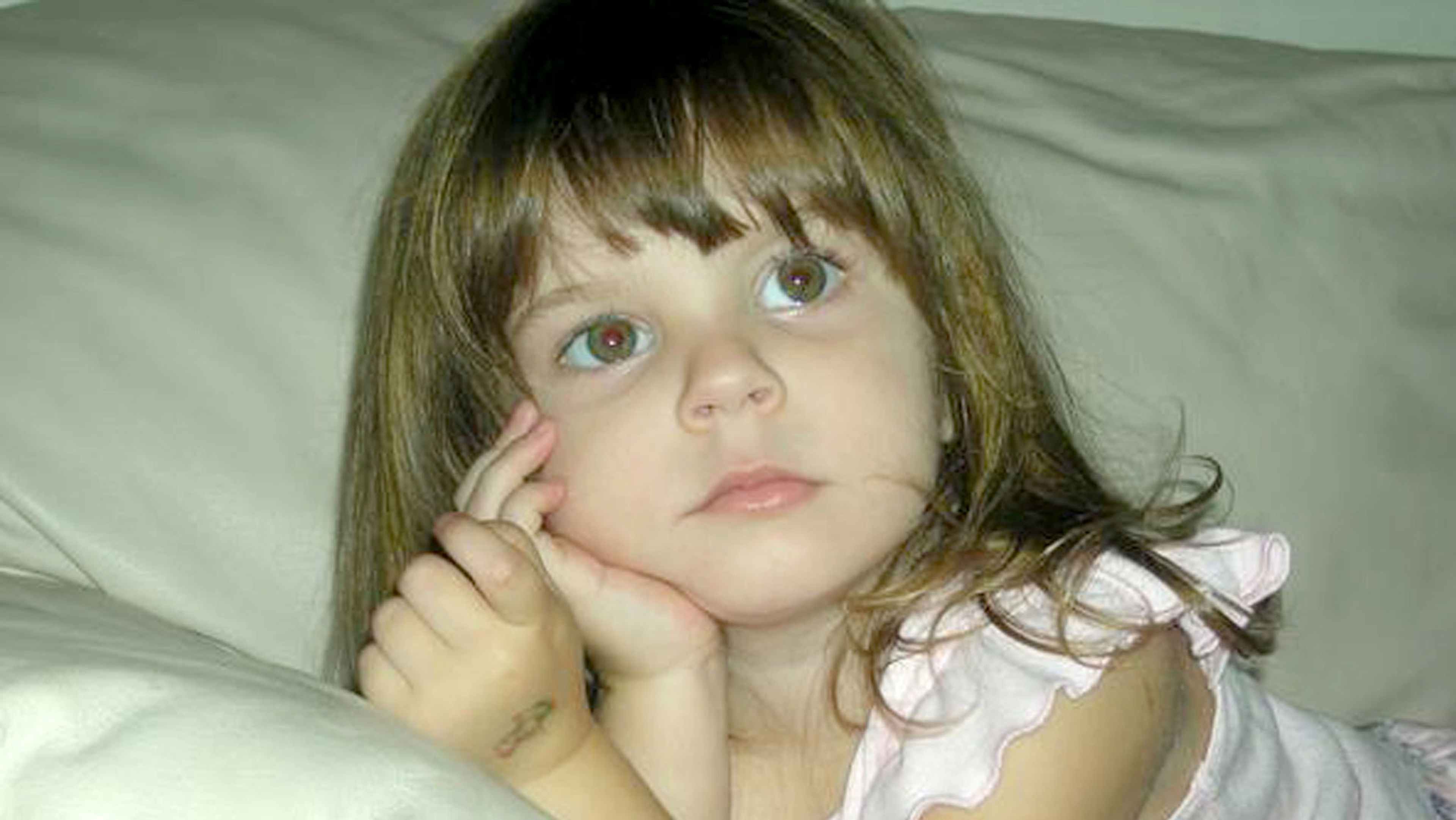A young girl with long, dark hair and blue eyes sits on a bed, her chin resting on her hands as she gazes thoughtfully.