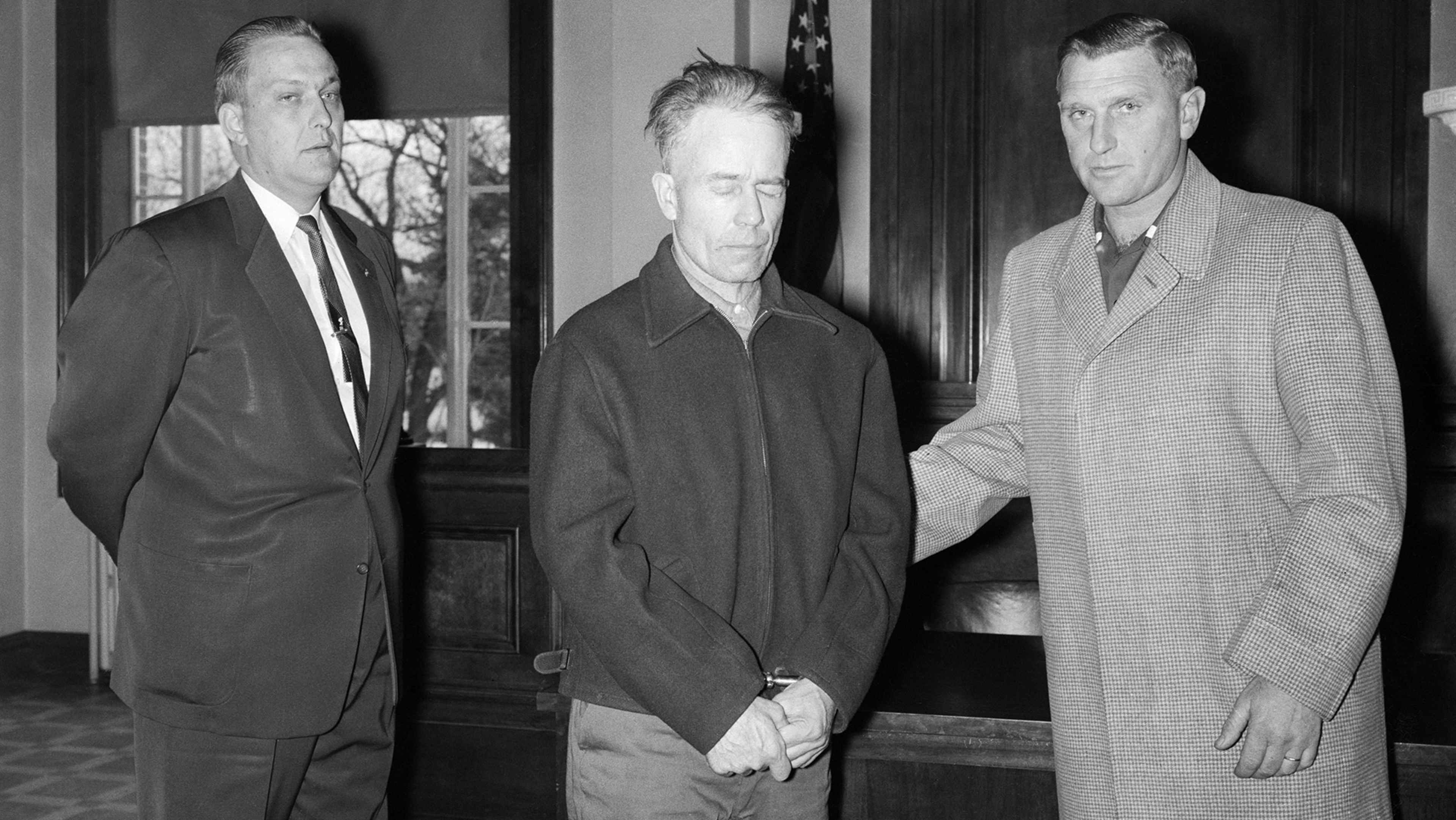 Three men in suits and coats standing in a doorway, with a window visible in the background.
