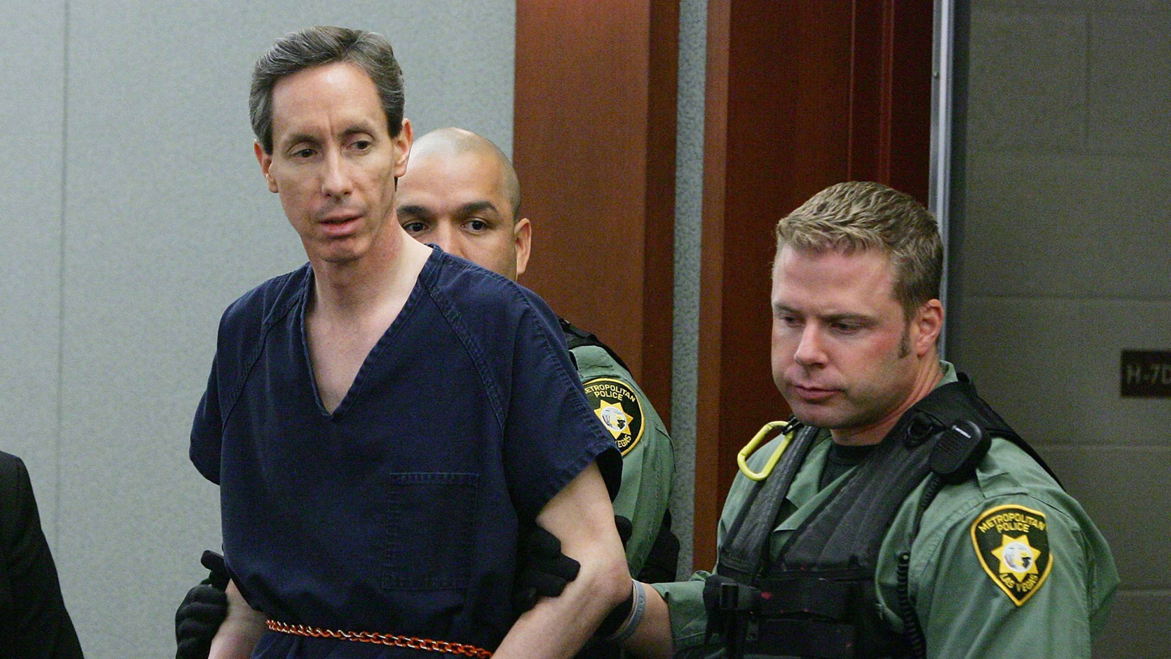 Two men in uniform, one wearing a navy blue shirt and the other wearing a green uniform, are standing in a room with a wooden door visible in the background.
